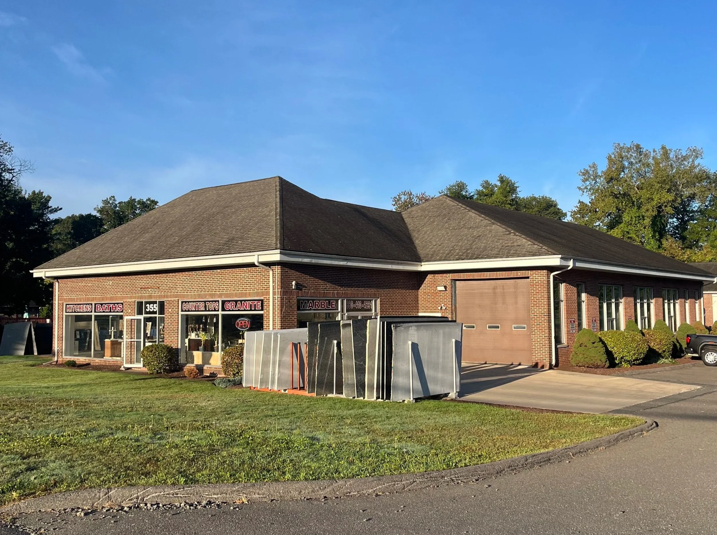 A brick building with large windows displays signs for countertops and granite with an open sign. Metal sheets are stacked outside near the entrance, and a parking lot with a truck is visible to the right. The building is surrounded by a green lawn and trees in the background under a clear blue sky.