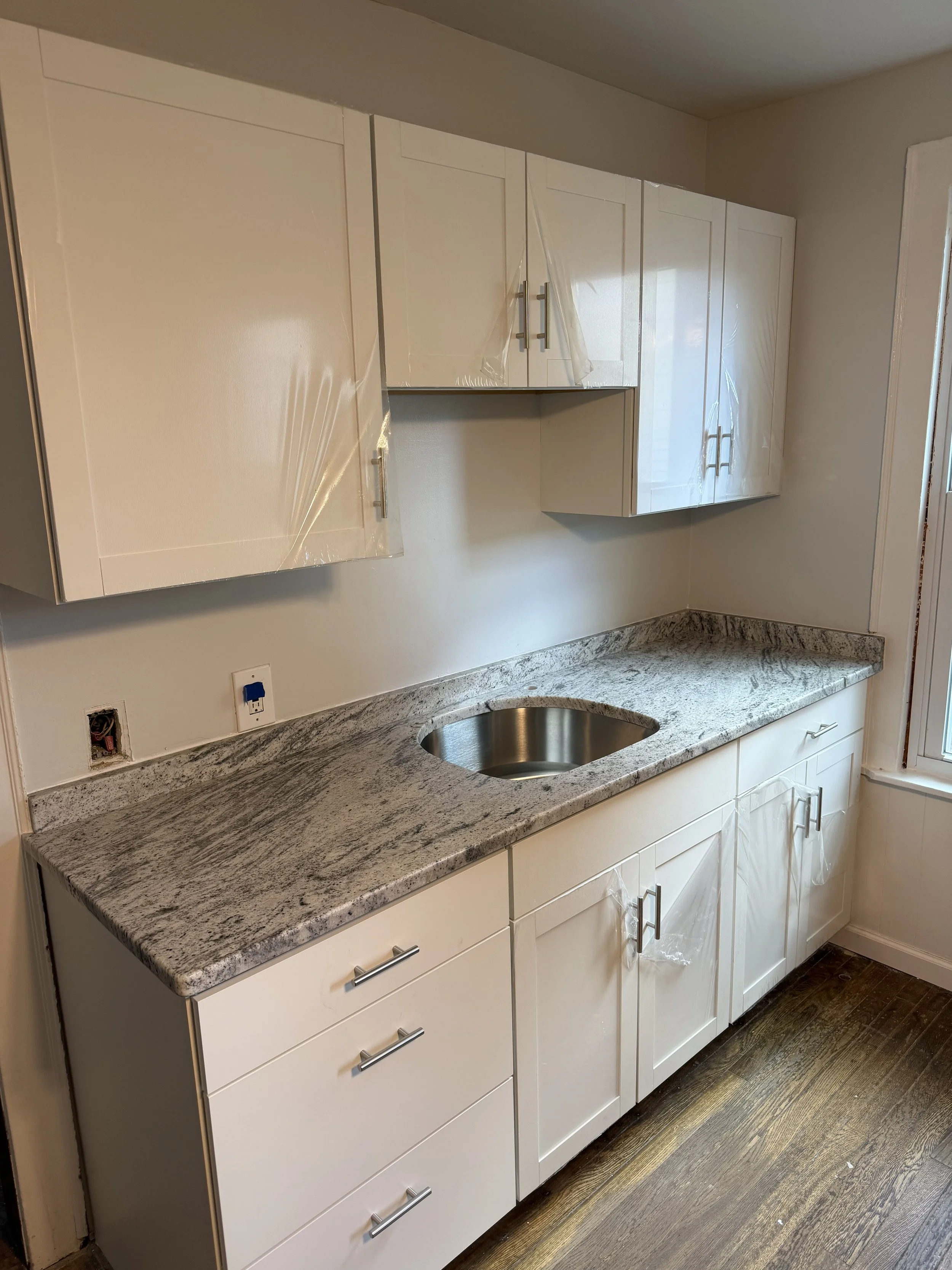 Kitchen with white cabinets, granite countertop, and stainless steel sink, under construction or renovation with some cabinets covered in plastic.