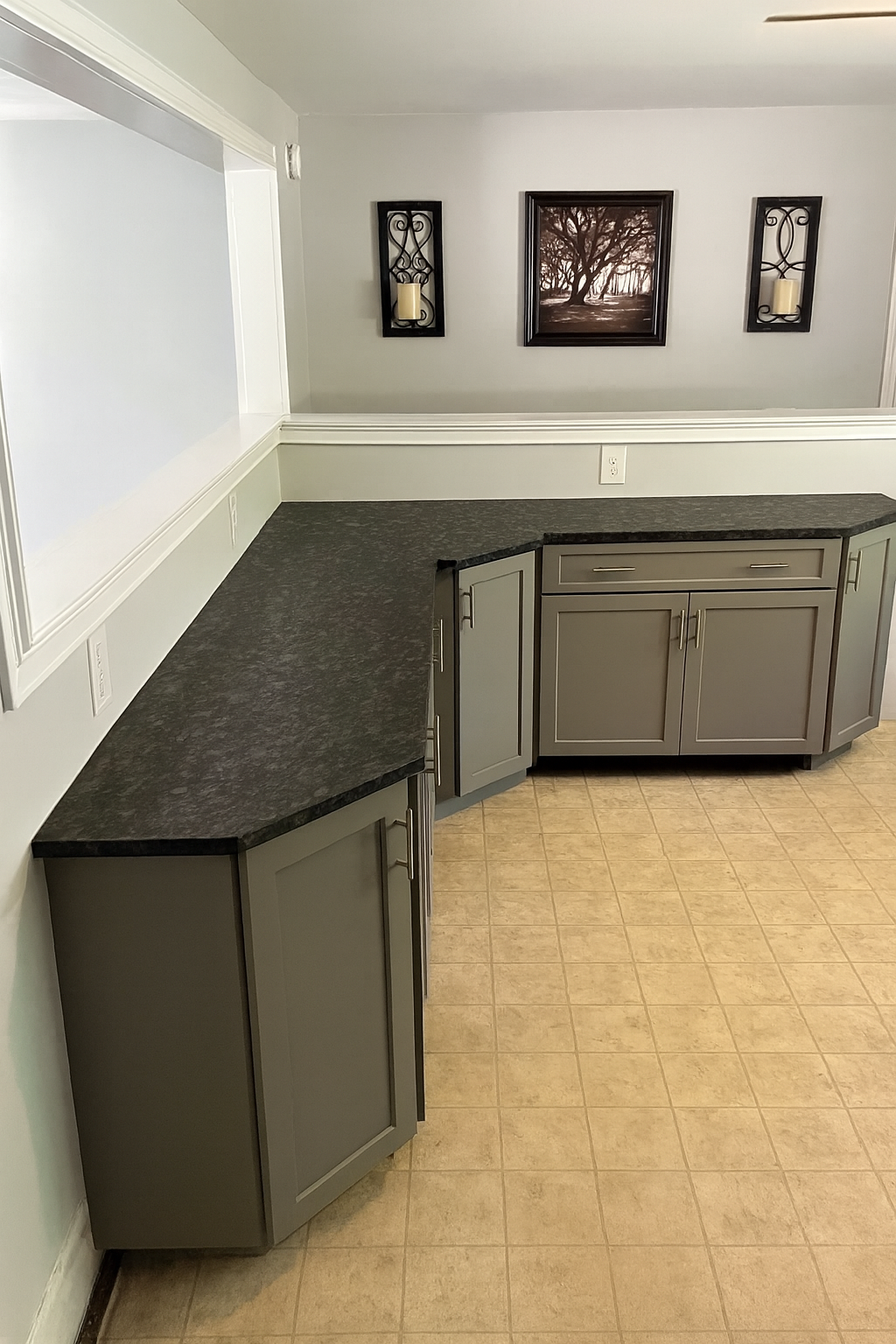 Kitchen corner with grey cabinets, black countertop, framed picture of a tree, and wall sconces with candles.