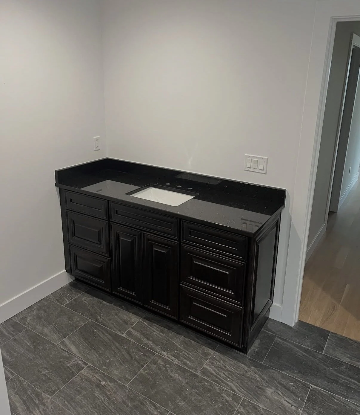 Black kitchen cabinet with a black countertop and a cutout for a sink, situated in a room with gray tiled flooring and white walls.