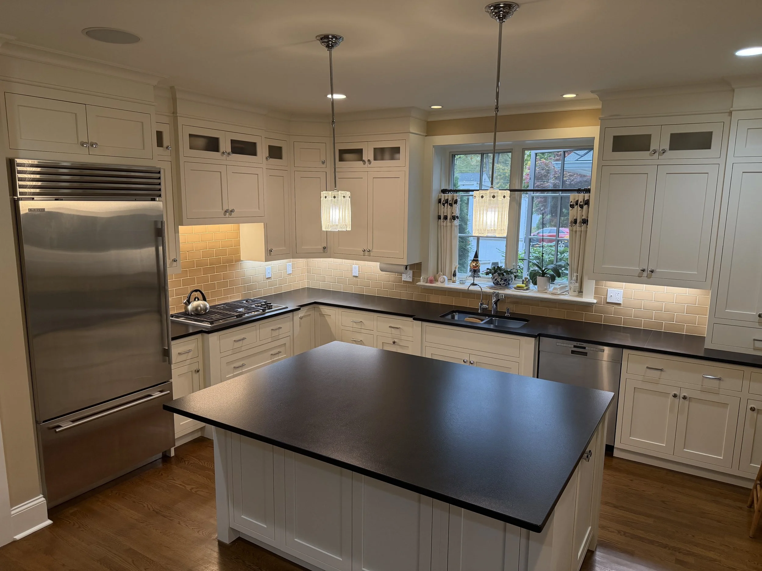 Bright kitchen with white cabinetry, black countertops, and a centered island. Stainless steel refrigerator, dishwasher, and a stove with a kettle. A window above the sink with curtains and kitchen plants. Pendant lights hanging over the island and recessed ceiling lights.