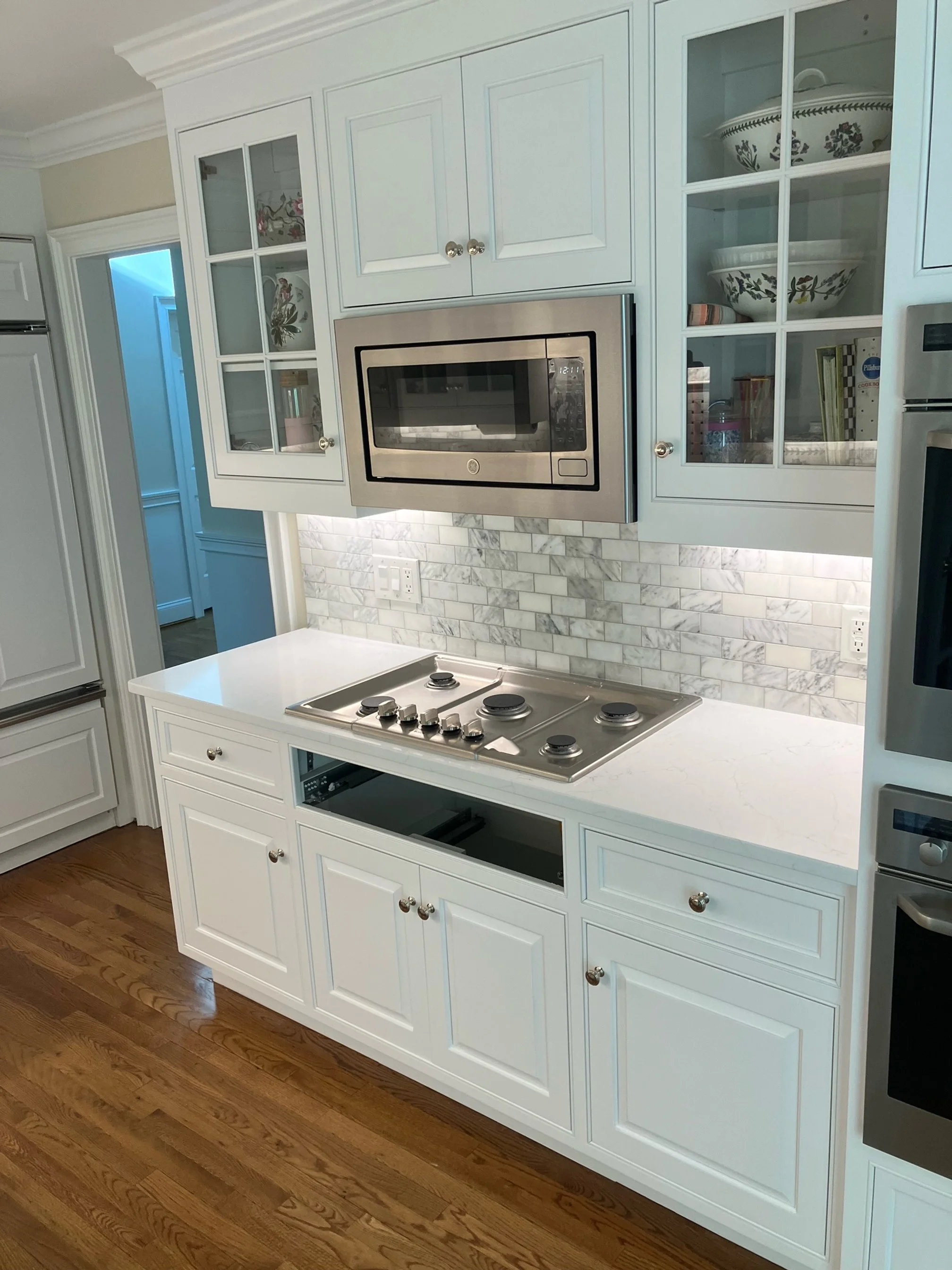White kitchen cabinetry with glass-paneled upper cabinets, a marble backsplash, built-in microwave, stovetop, and hardwood flooring.