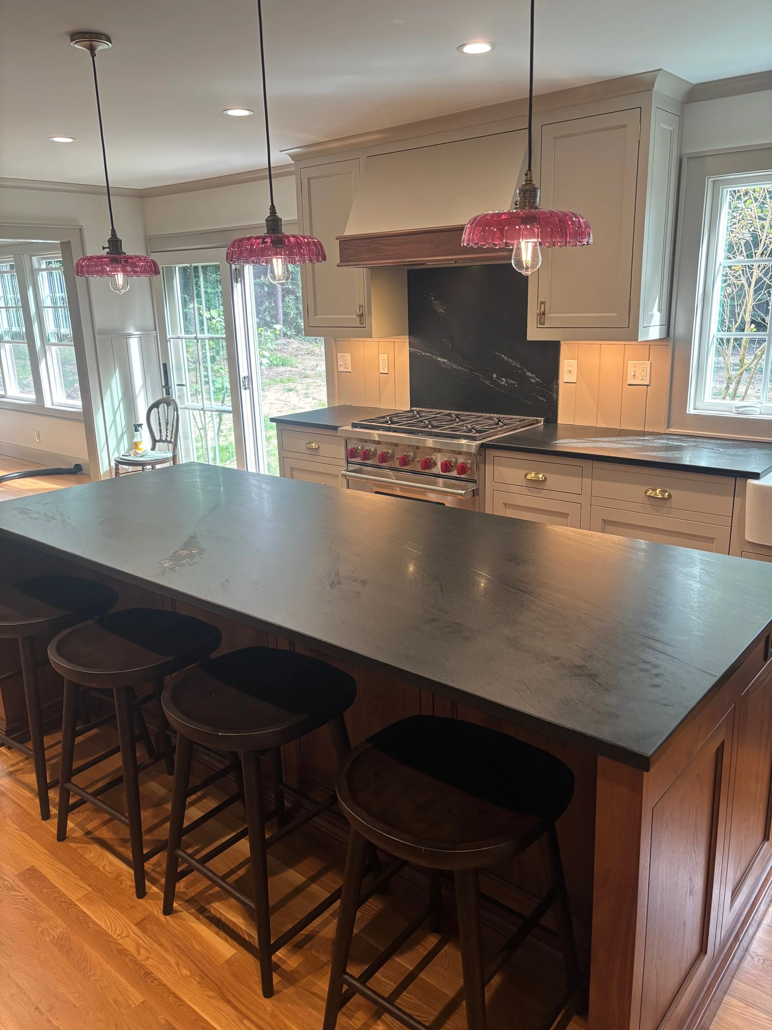 Kitchen island with four black stools, gas stove with red knobs, pendant lights with pink glass shades, and windows allowing natural light.