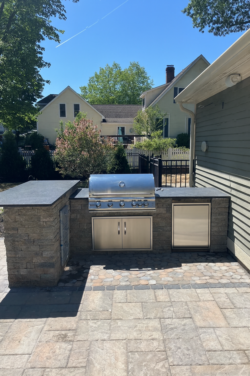 Outdoor patio with built-in grill, counter space, and storage cabinets, surrounded by a stone and brick wall, with houses and trees in the background under a clear blue sky.