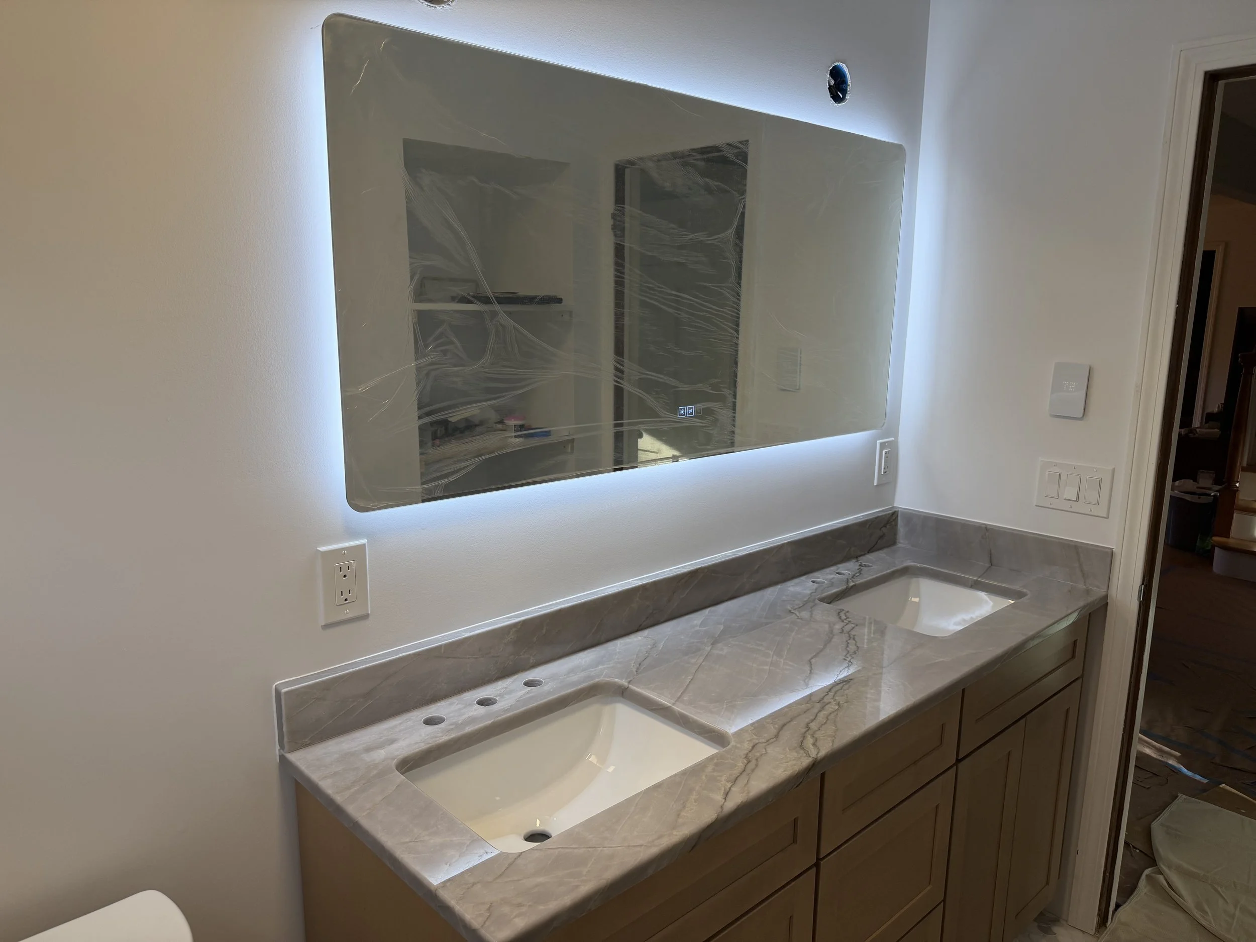 Bathroom vanity with a marble countertop and two inset sinks, large framed mirror with LED lighting, white walls, electrical outlets, and unfinished wall above the mirror.