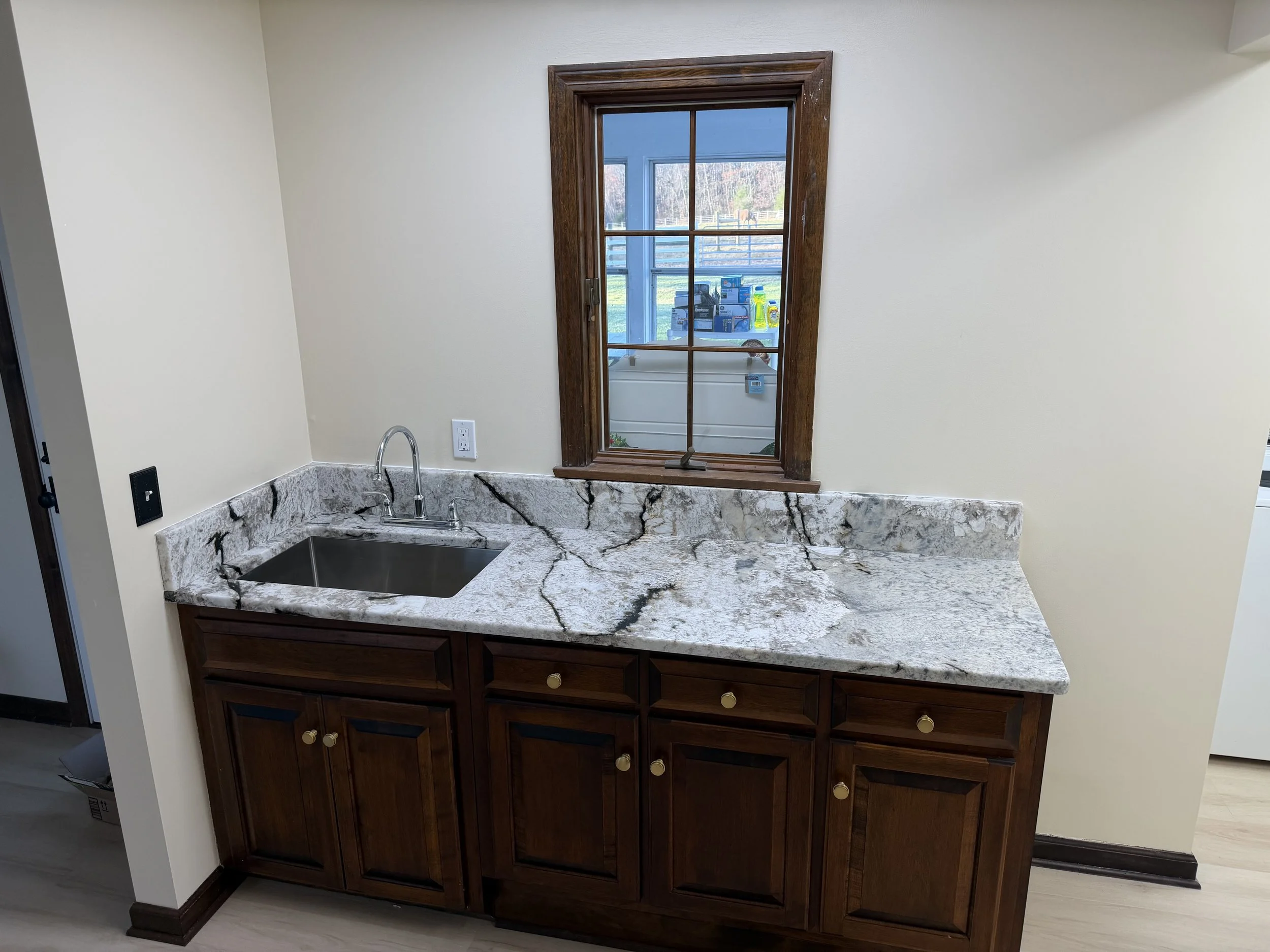 Kitchen sink with marble countertop, wooden cabinets, and a window above the sink showing an outside view with boxes and trees.