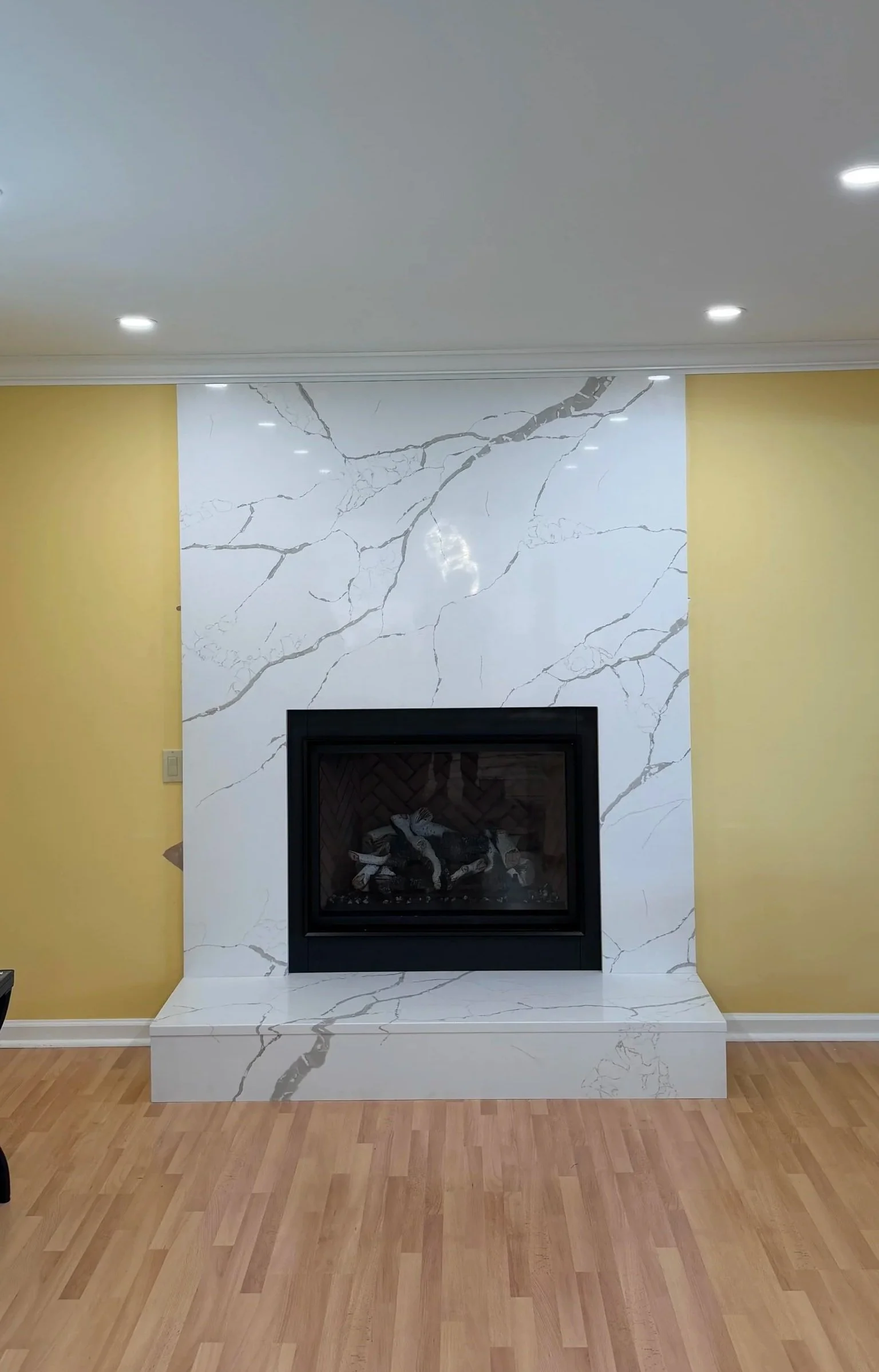 Living room fireplace with white marble tiles with gray veining, yellow walls on either side, wood flooring, ceiling lights, and a black enclosed fire.