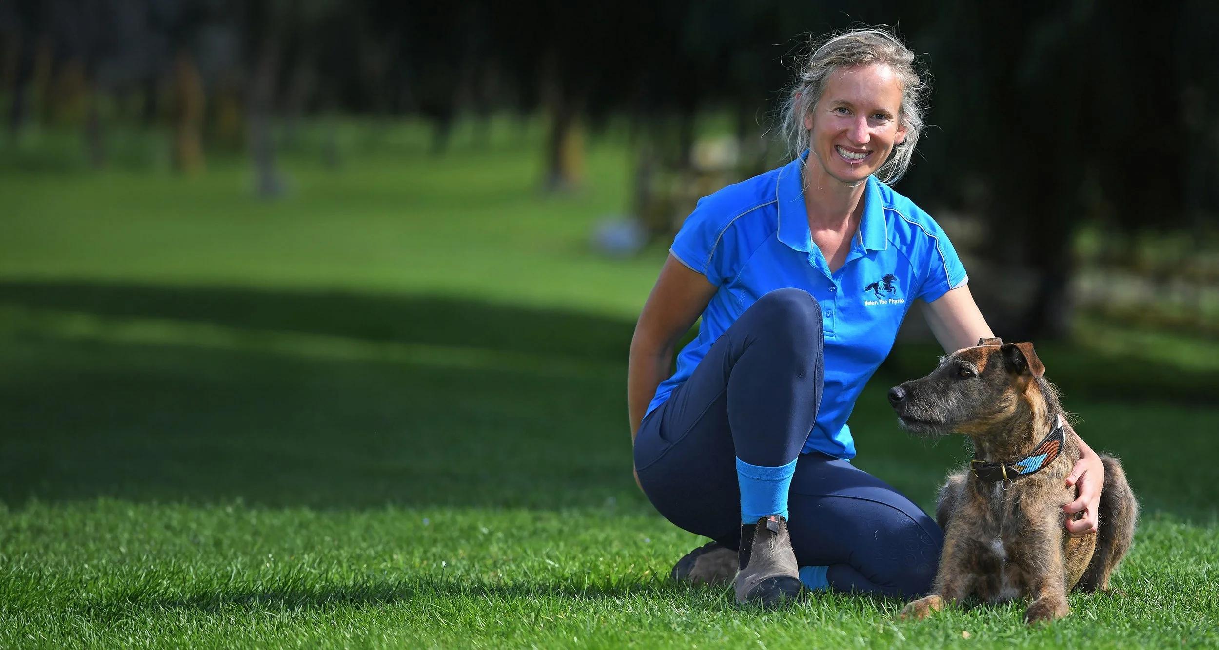 Helen the Physio crouching next to her dog Rhubarb in an open green field