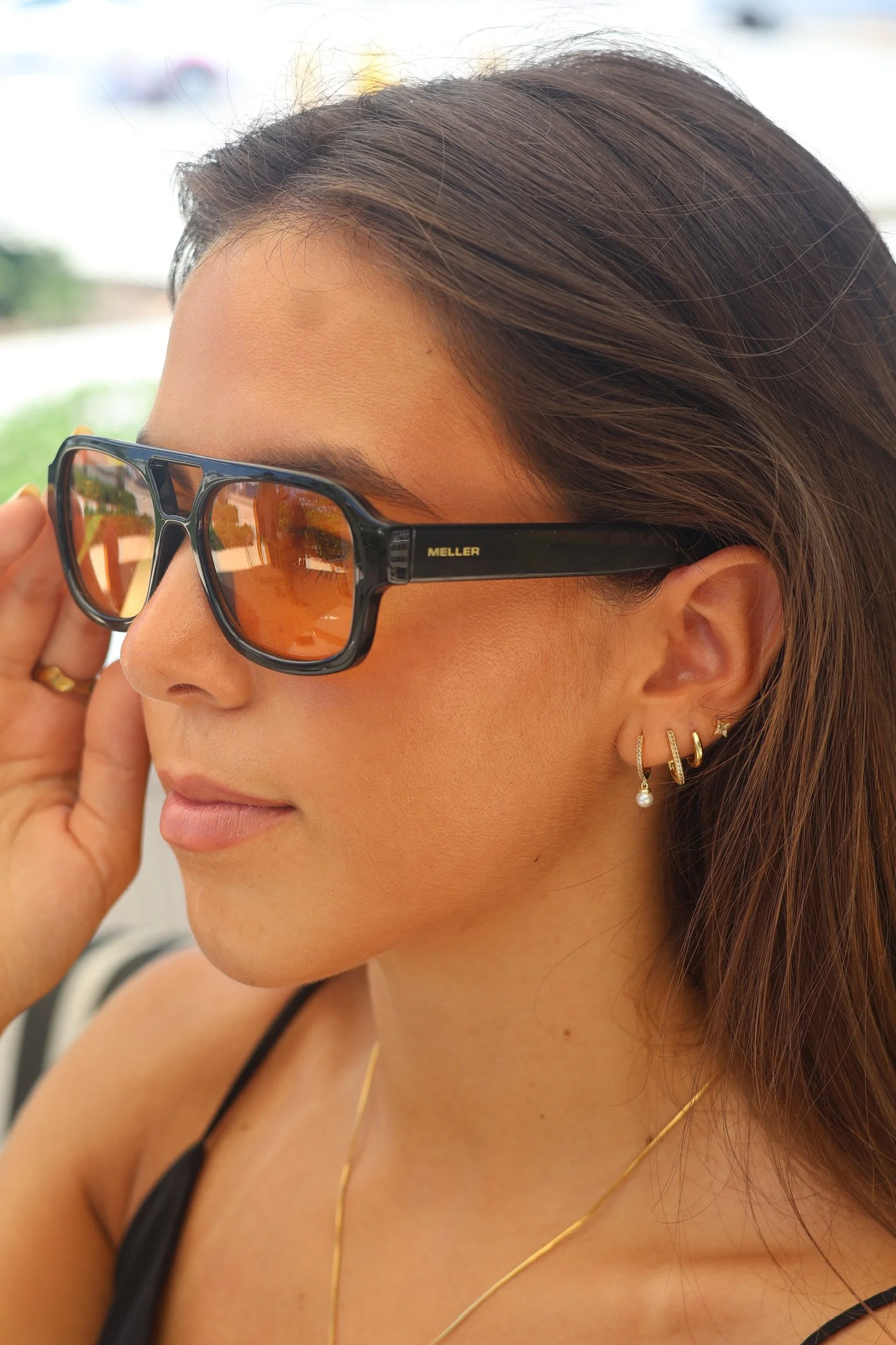 Close-up of a woman wearing black sunglasses with orange lenses, gold jewelry including earrings and a necklace, and a black top.