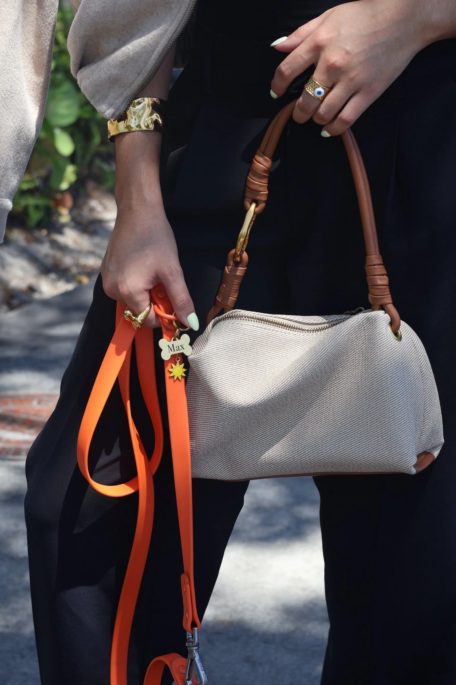 A person's hand holding a leash with an orange strap, attached to a dog. The person is wearing a ring with a blue eye design, a gold bracelet, and has painted nails. They are dressed in dark pants and a beige jacket with gold cuffs. There is a beige bag with brown handles hanging from their arm.