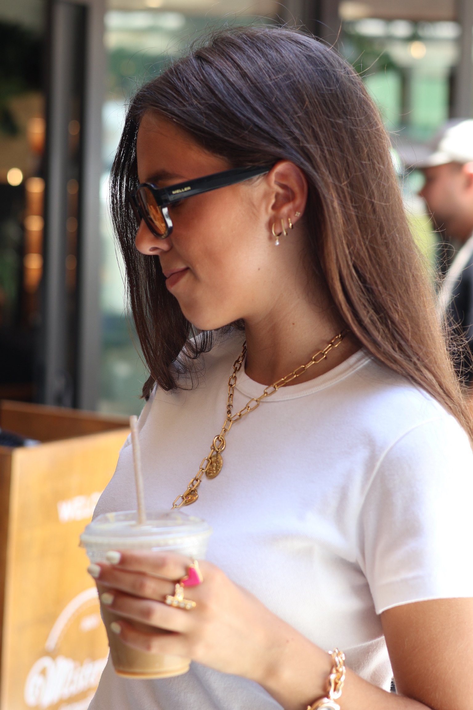 A young woman with long brown hair, wearing black sunglasses, gold jewelry including earrings, a necklace, and a bracelet, is holding a cold coffee drink with a straw. She is wearing a white t-shirt and appears to be in a cafe or cafe-like setting.