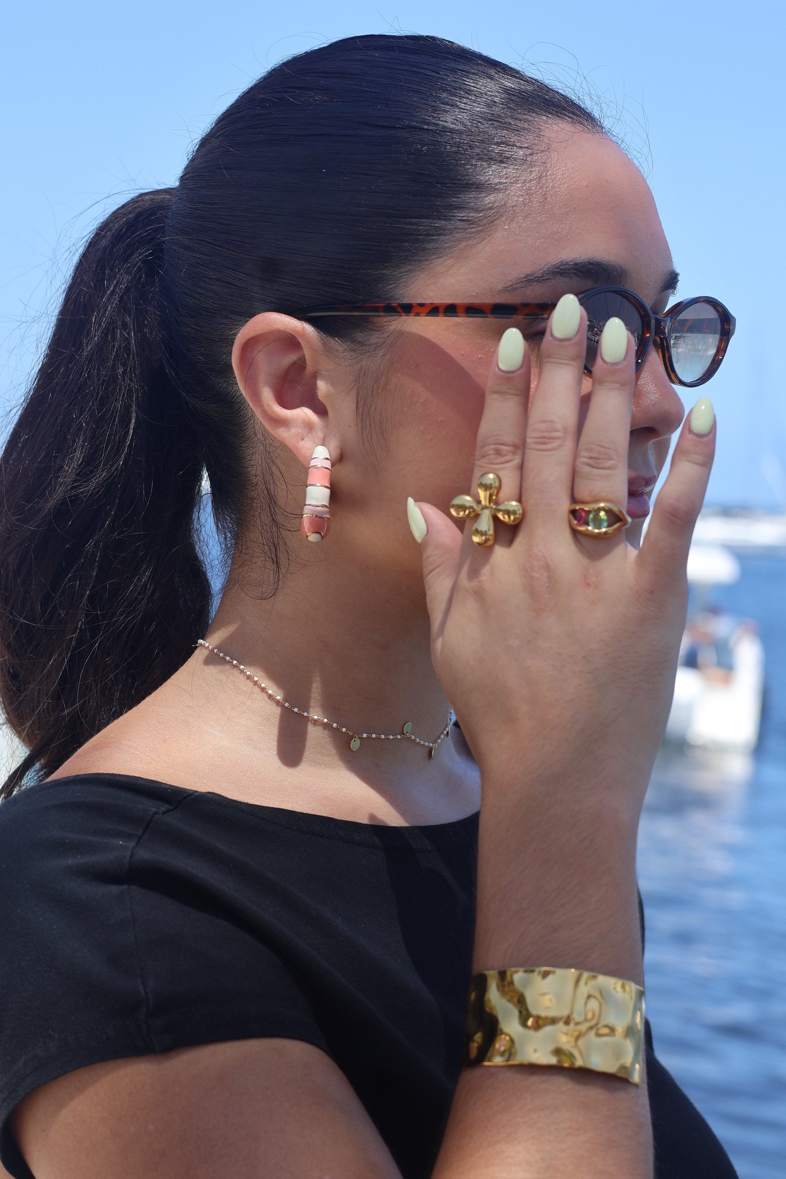 Close-up side profile of a woman with dark hair tied back, wearing tortoiseshell sunglasses, colorful striped earrings, and gold rings, standing outdoors by the water on a sunny day.