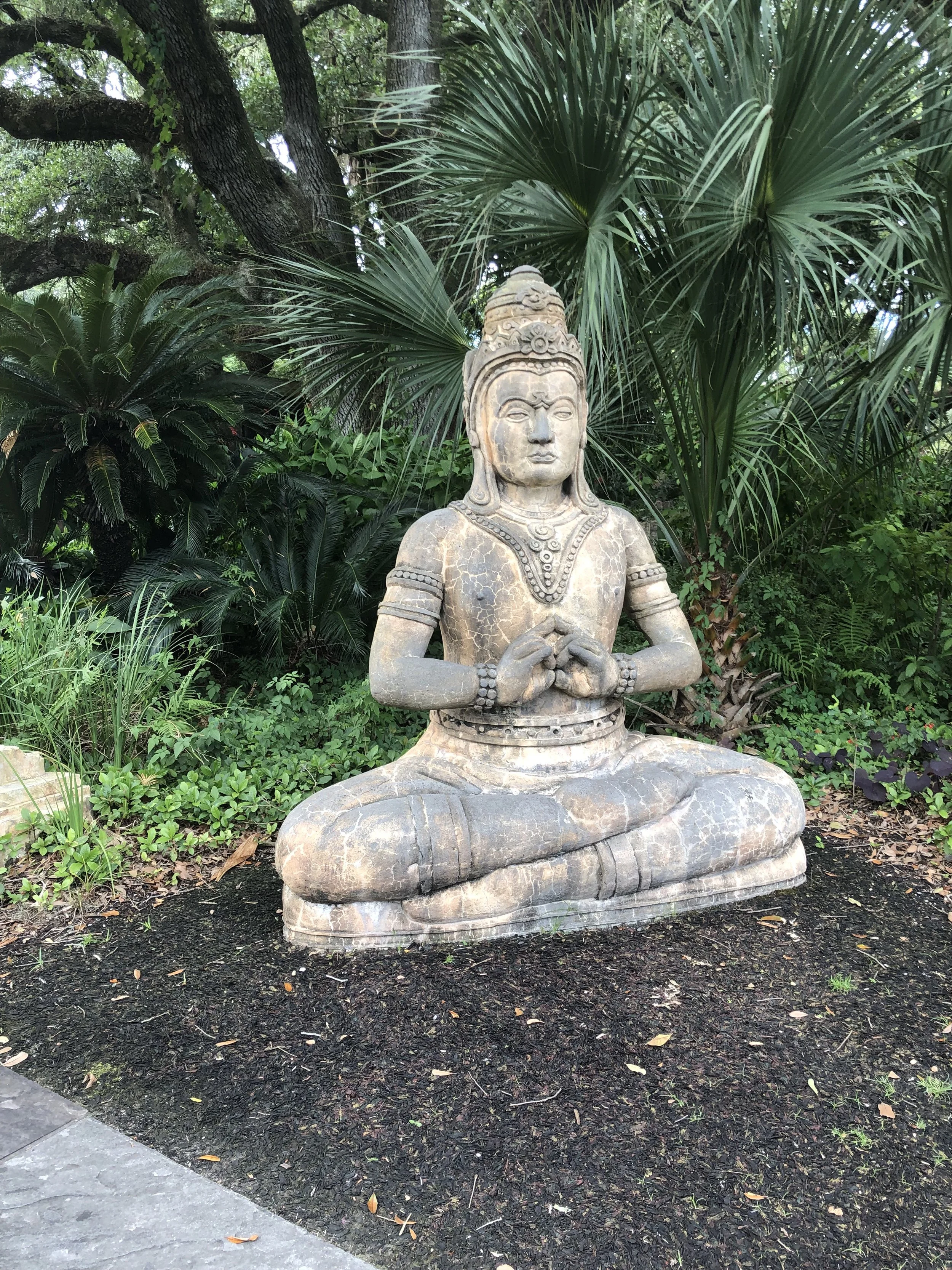A stone sculpture of a meditating figure with hands in a mudra, seated cross-legged on the ground, surrounded by lush green plants and trees.