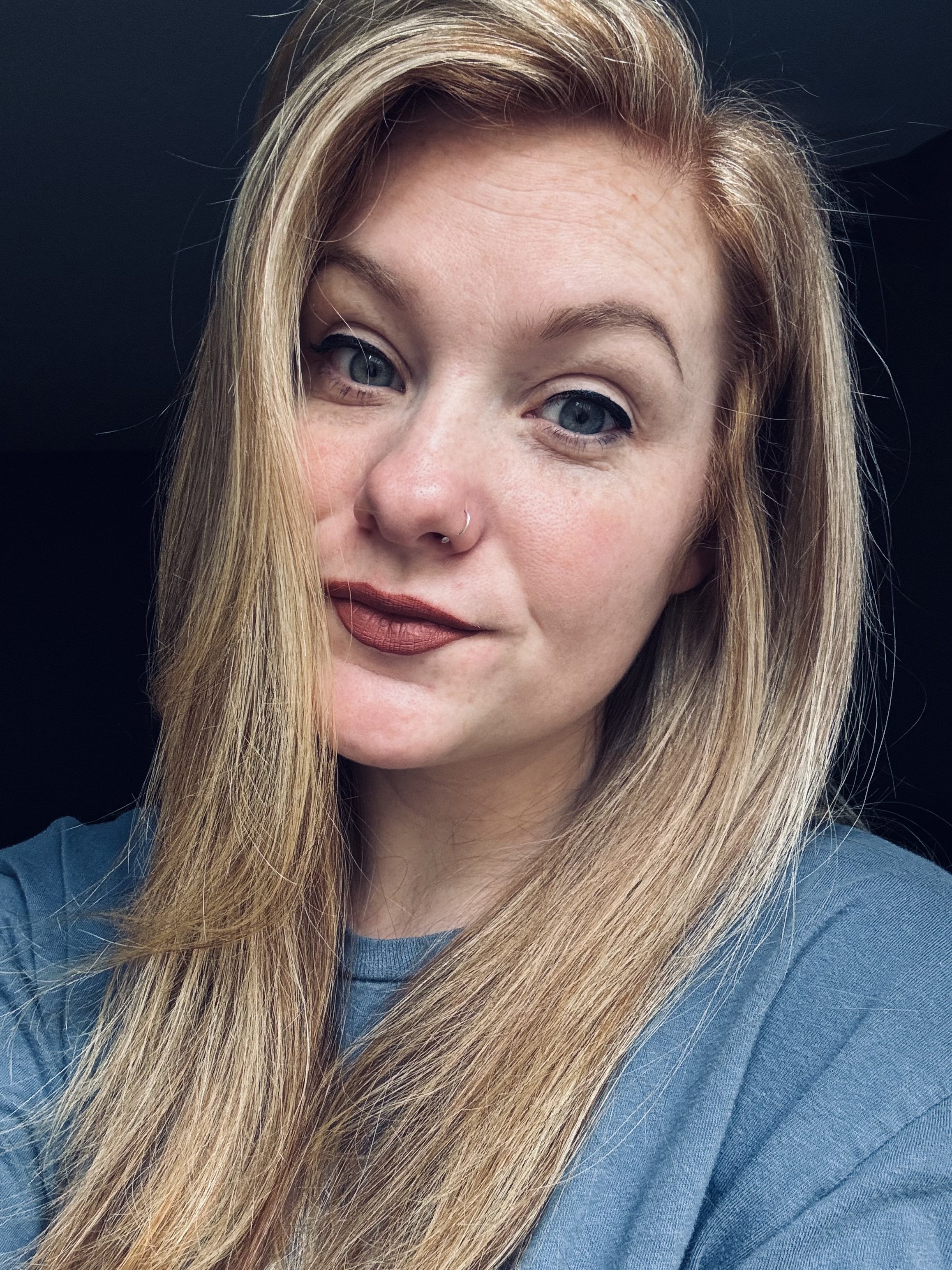 A young woman with long blonde hair, blue eyes, and a nose ring, wearing a blue top, looking at the camera with one eye slightly closed, against a dark background.