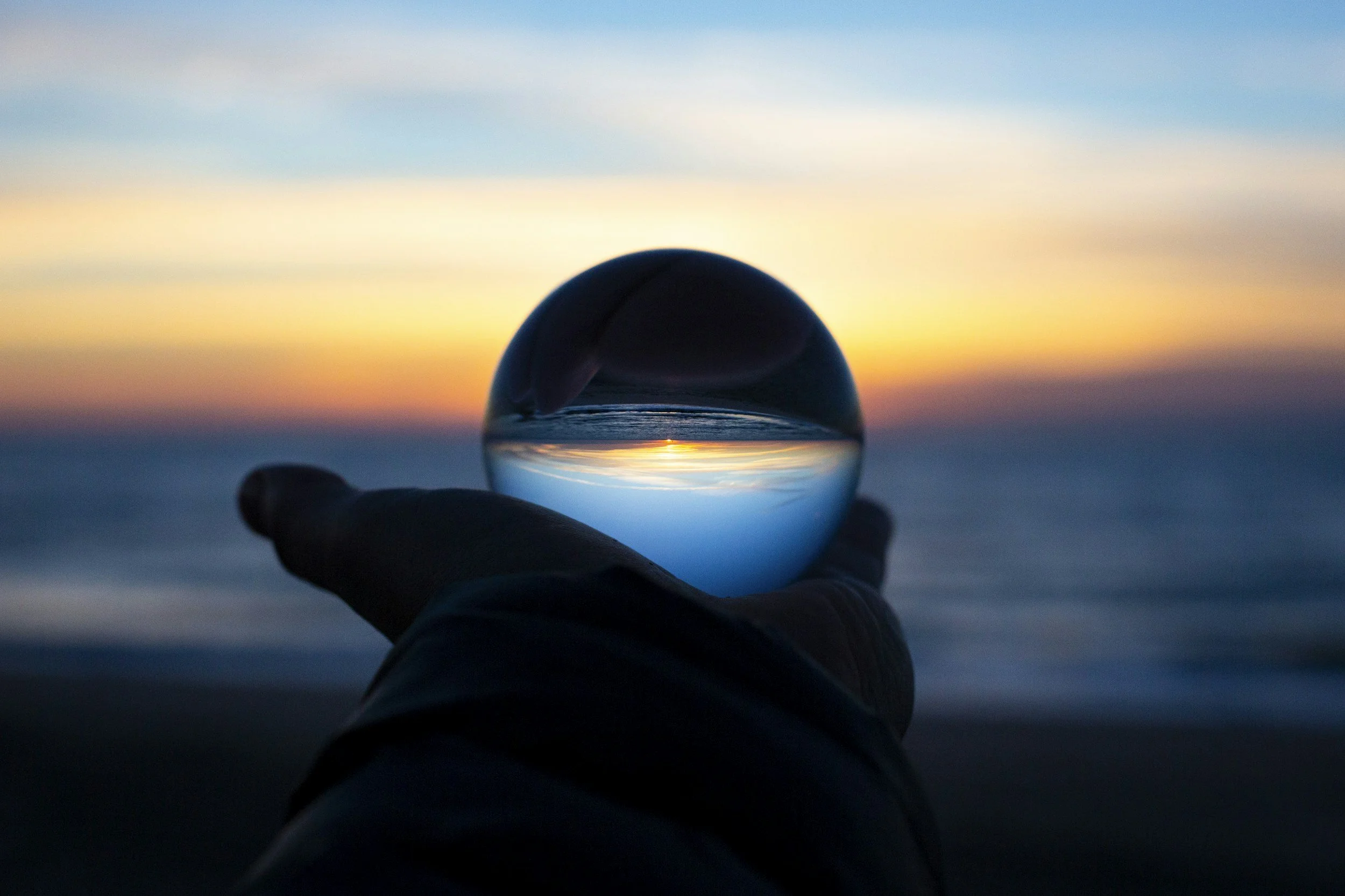 Hand holding glass sphere reflecting sunset over ocean horizon.