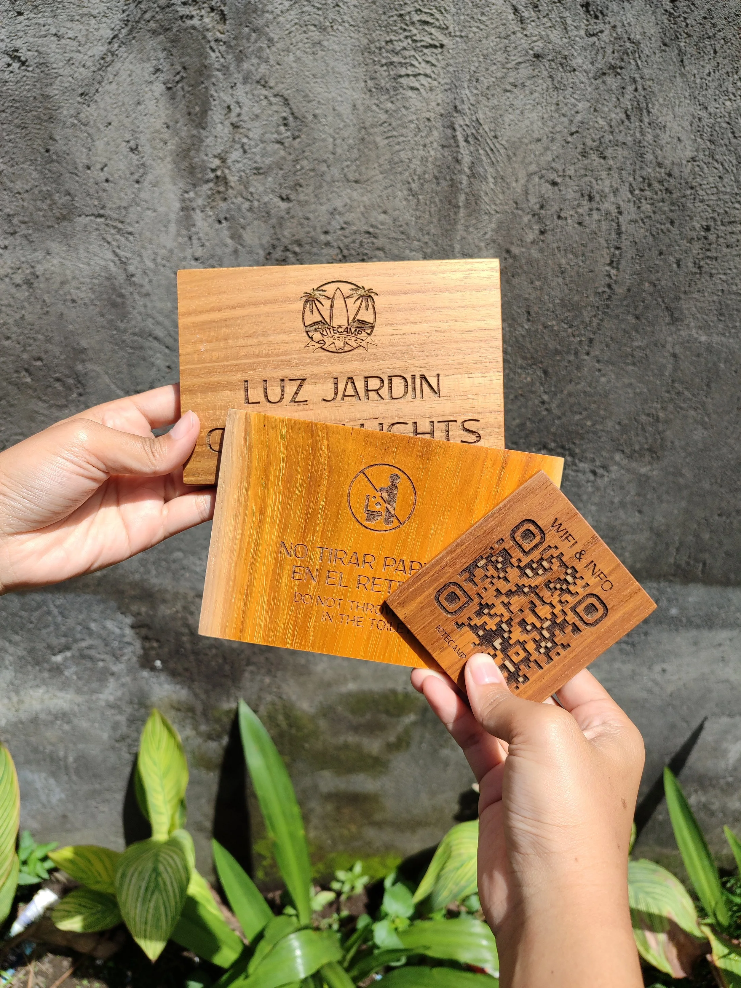 Person holding three wooden signs and a QR code sign in front of a stone wall and green plants. The signs include information about garden lights, no littering, and Wi-Fi QR code.