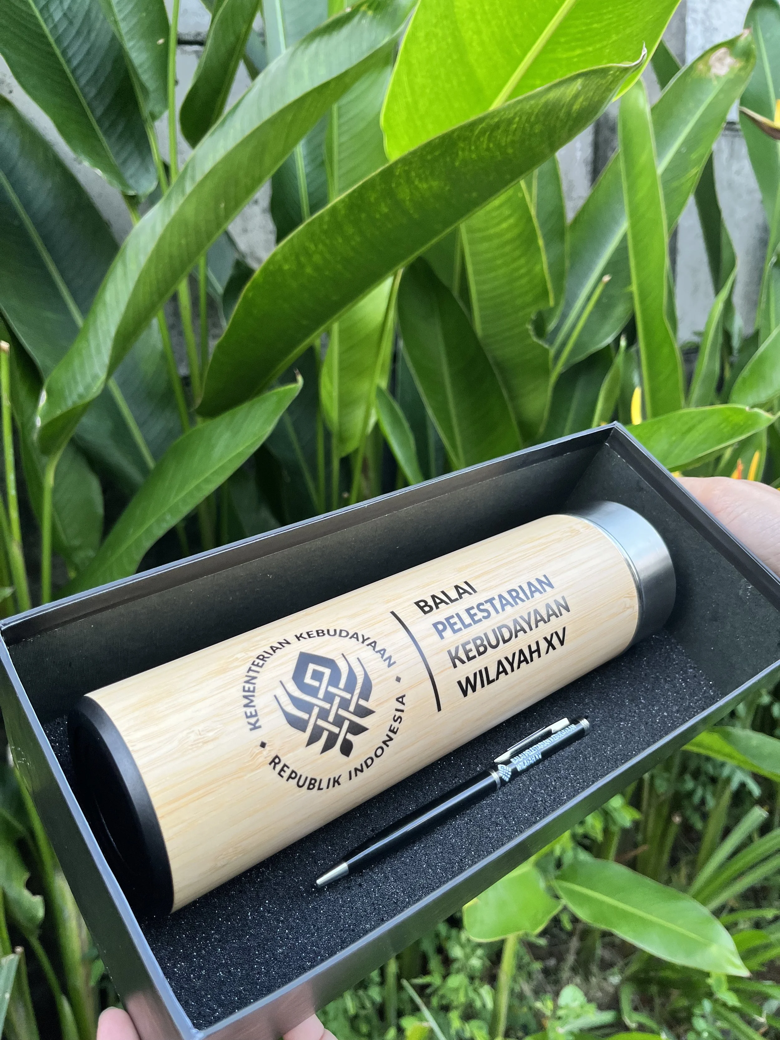 A wooden commemorative cylinder with Indonesian text and logo, resting in a black box with foam padding, accompanied by a black pen, held in front of lush green foliage.