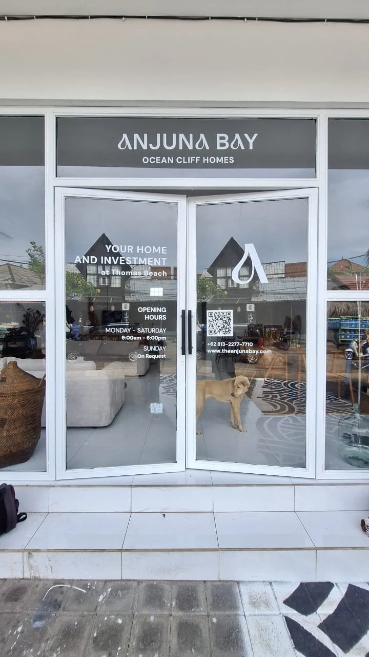 Storefront of Anjuna Bay Ocean Cliff Homes, with glass doors, a Labrador retriever inside, and information about opening hours and contact details