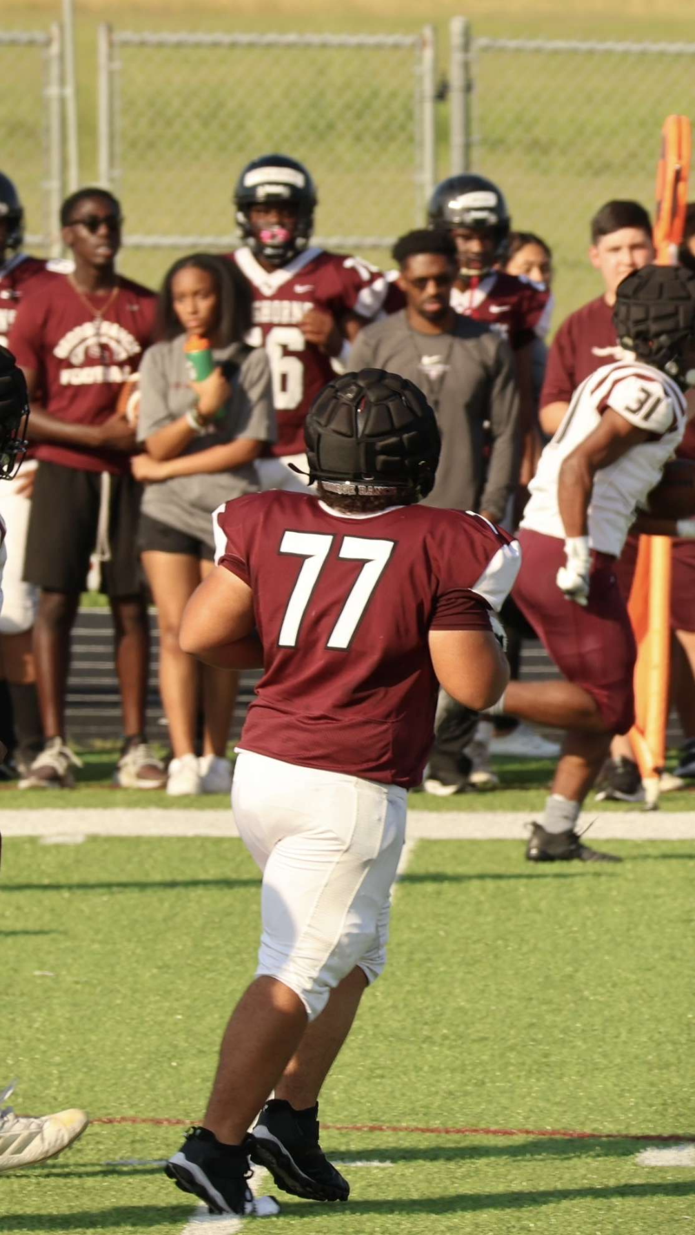 American football player wearing jersey number 77 in maroon and white, running on the field during a game with spectators and teammates in the background.