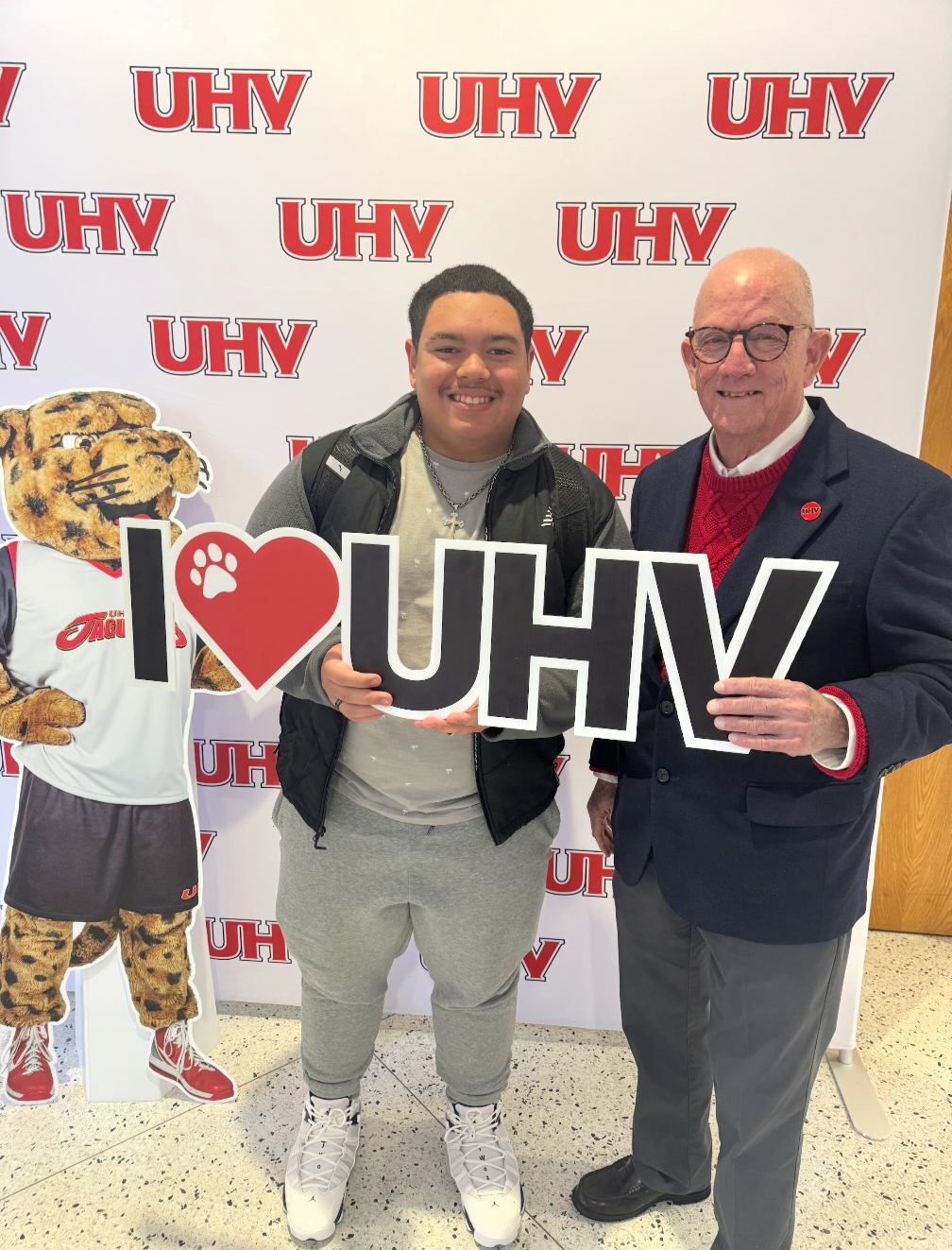 Two men standing together, holding a sign that says 'I love UHV', in front of a UHV branded step and repeat backdrop. One man is younger, wearing a gray hoodie and light gray sweatpants, and the older man is dressed in a dark blazer, red sweater, and glasses. A UHV mascot, a cheetah, stands on the left, wearing a UHV sports jersey and shorts.