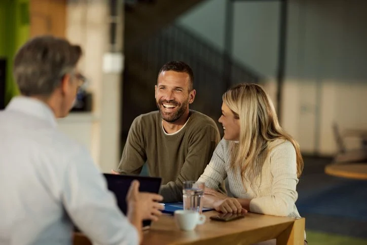 Deux jeunes adultes souriants discutent avec un homme plus âgé dans un café ou un restaurant.