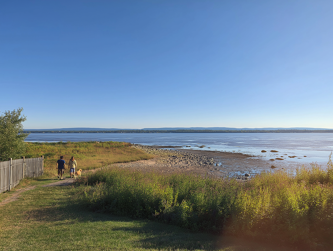 Deux personnes marchent avec un chien sur un sentier côtier, devant une plage rocheuse et un grand lac, sous un ciel clair.