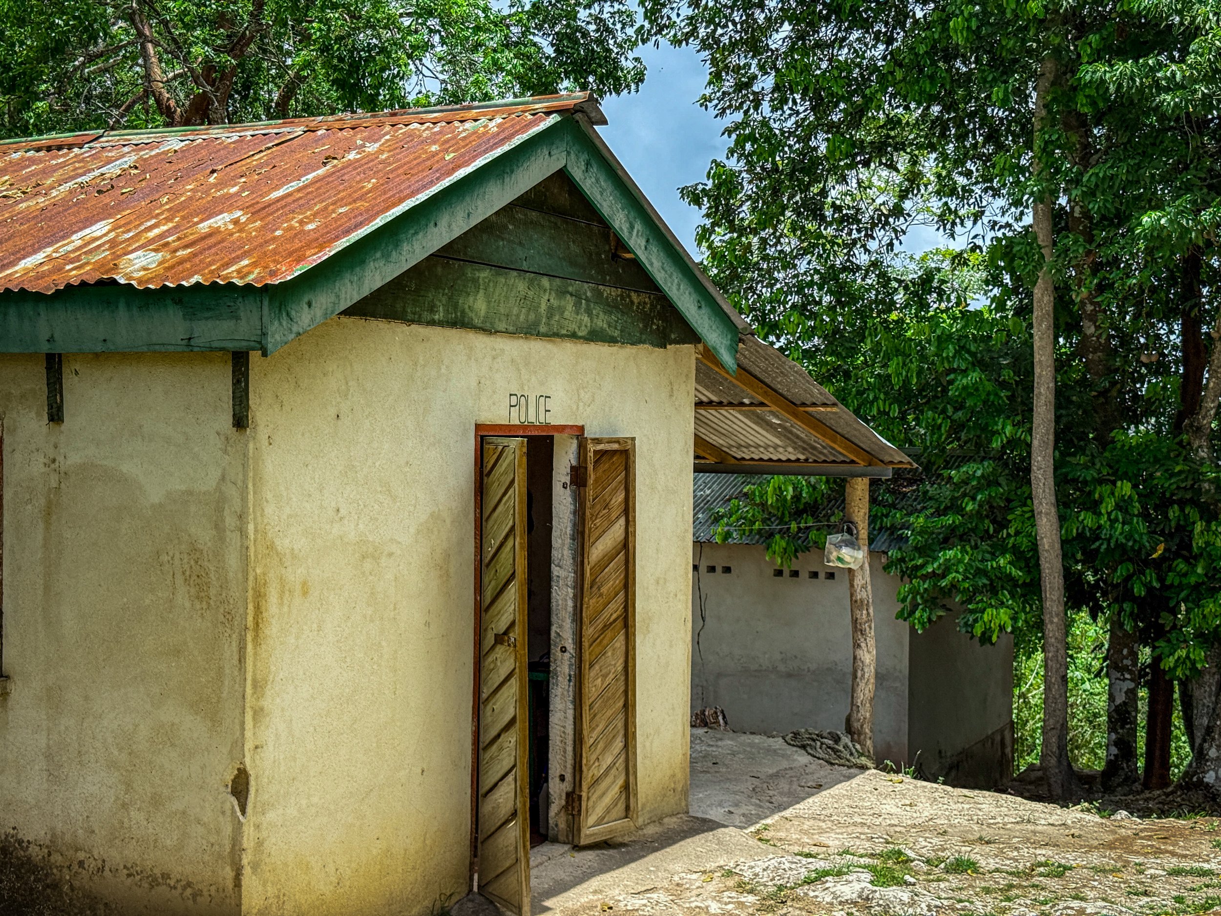 Small building with a rusty metal roof and open wooden door labeled "POLICE," surrounded by trees and greenery.