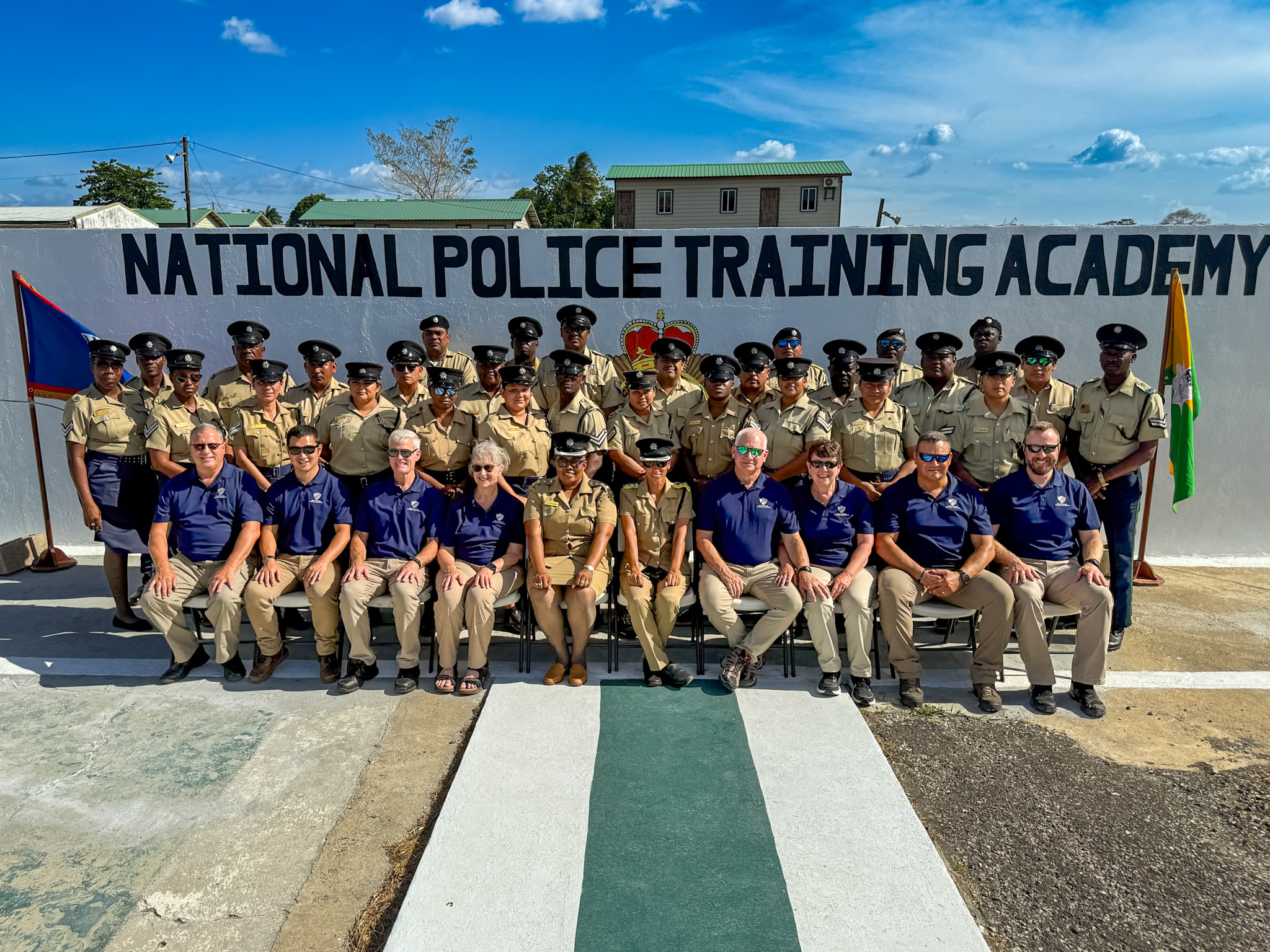 Group of police officers and staff posing in front of a wall with the text 'National Police Training Academy', with flags on either side, outdoors under a blue sky.