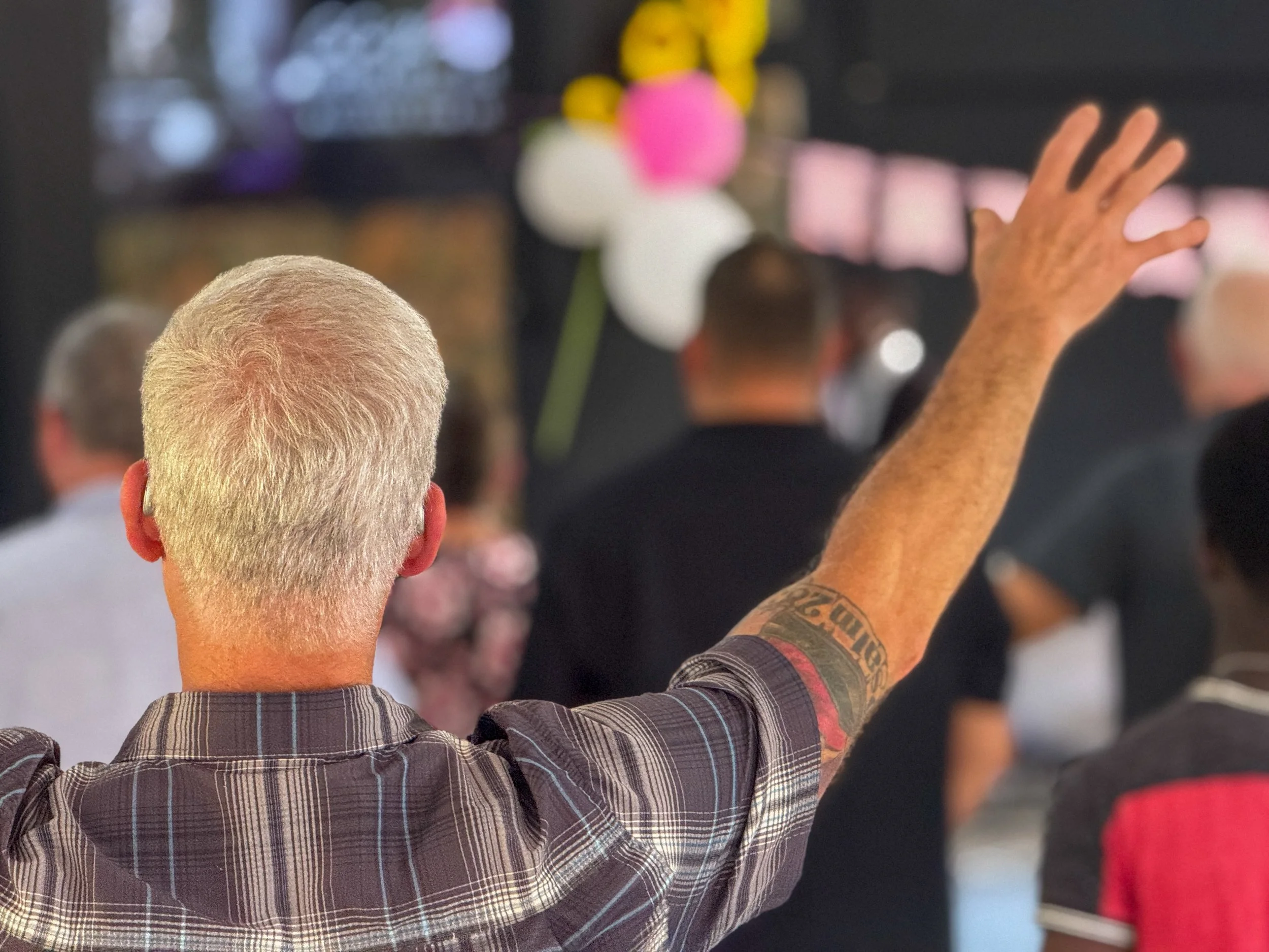A man with blond hair and a tattoo on his arm is raising his hand during a public event or gathering, with other people in the background.