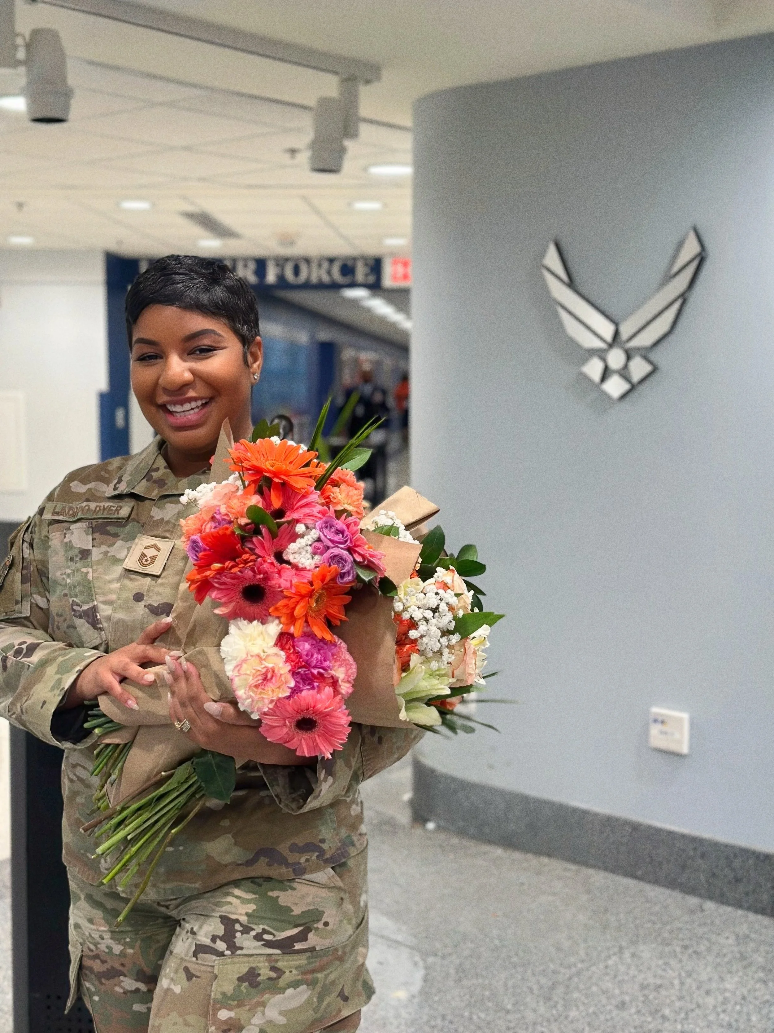 A smiling woman in military uniform holding a large bouquet of colorful flowers inside a building, with a military emblem on the wall behind her.