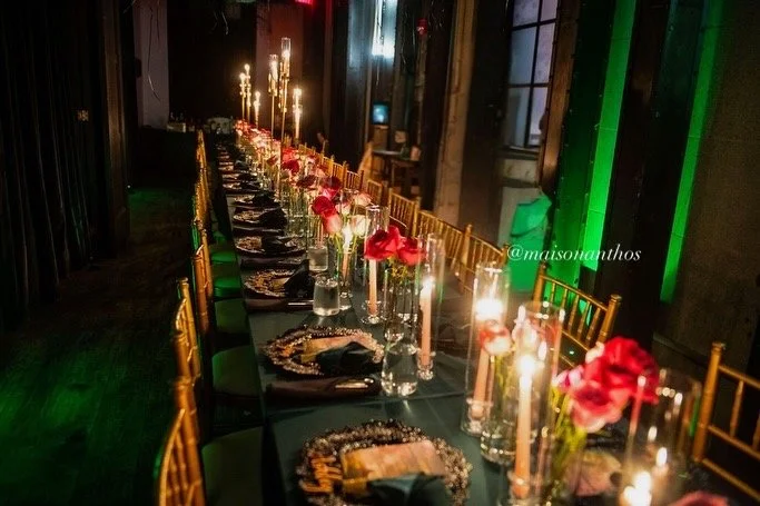 Long banquet table decorated with candles, red flowers, and ornate chargers, set for a formal event in a dimly lit room with dark walls and green accent lighting.