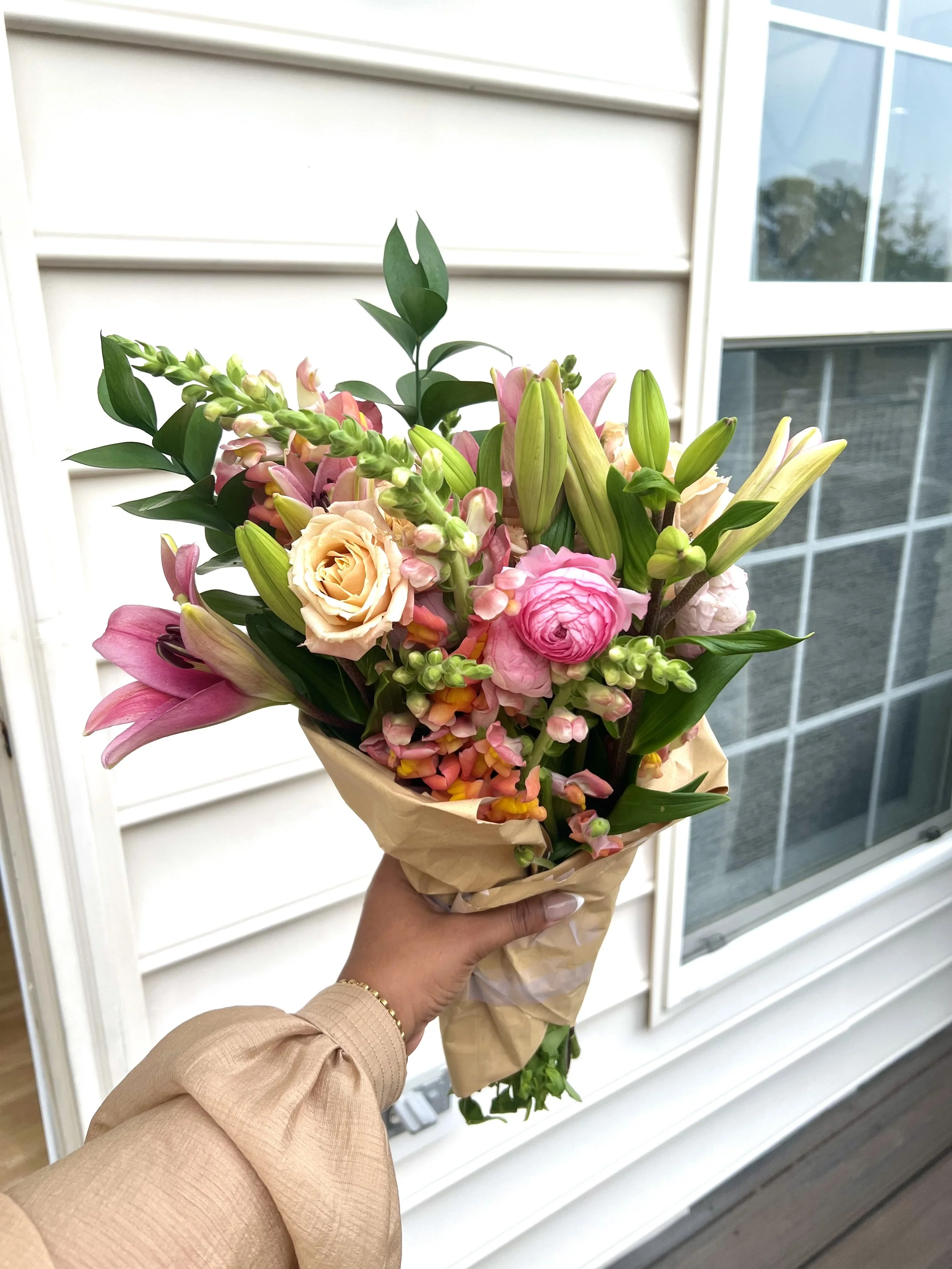Hand holding a bouquet of pink, peach, and white flowers with green leaves outside a house with white siding and window.