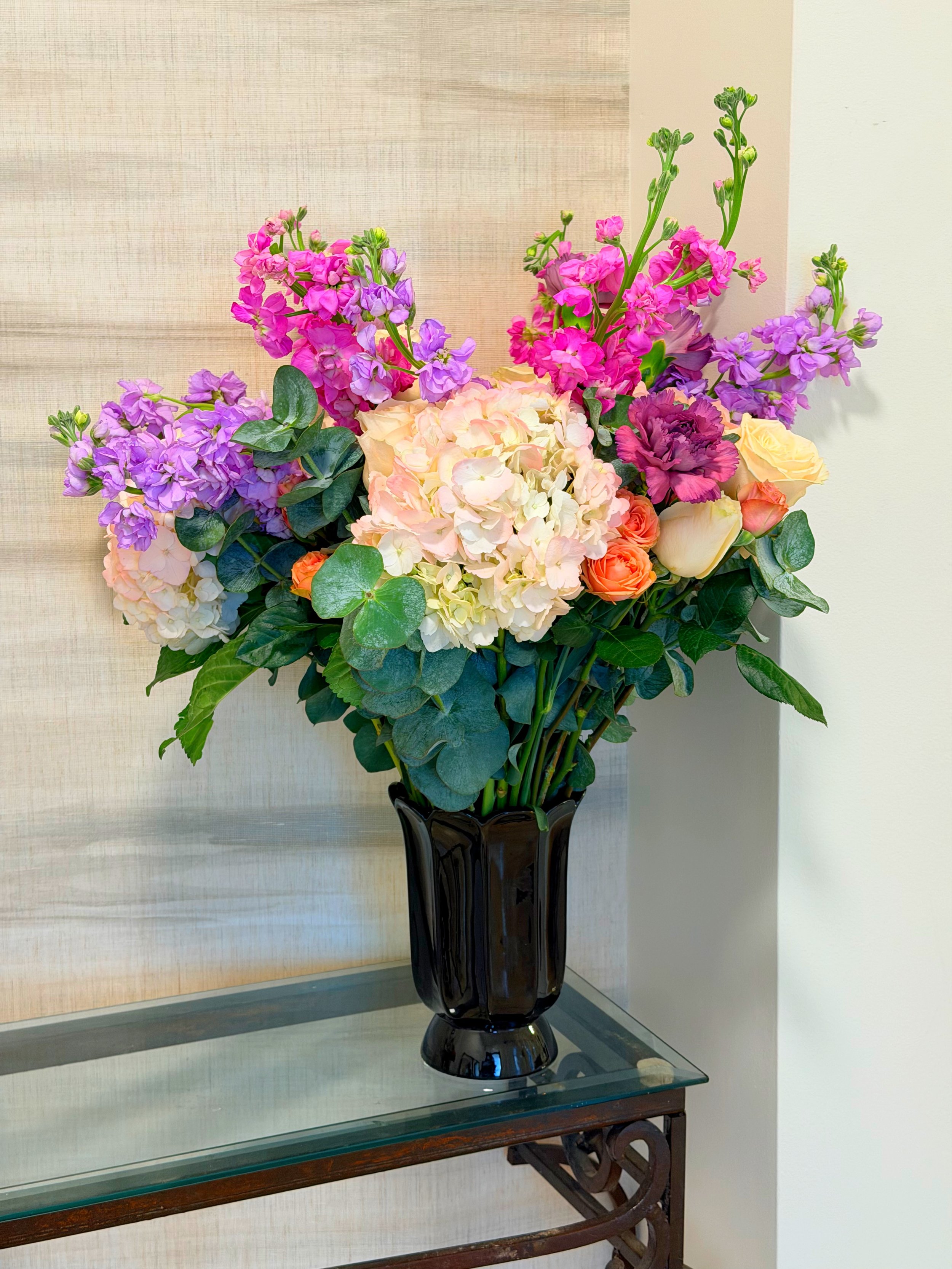 A black vase with a bouquet of pink, purple, white, and orange flowers on a glass-topped table against a light-colored wall.