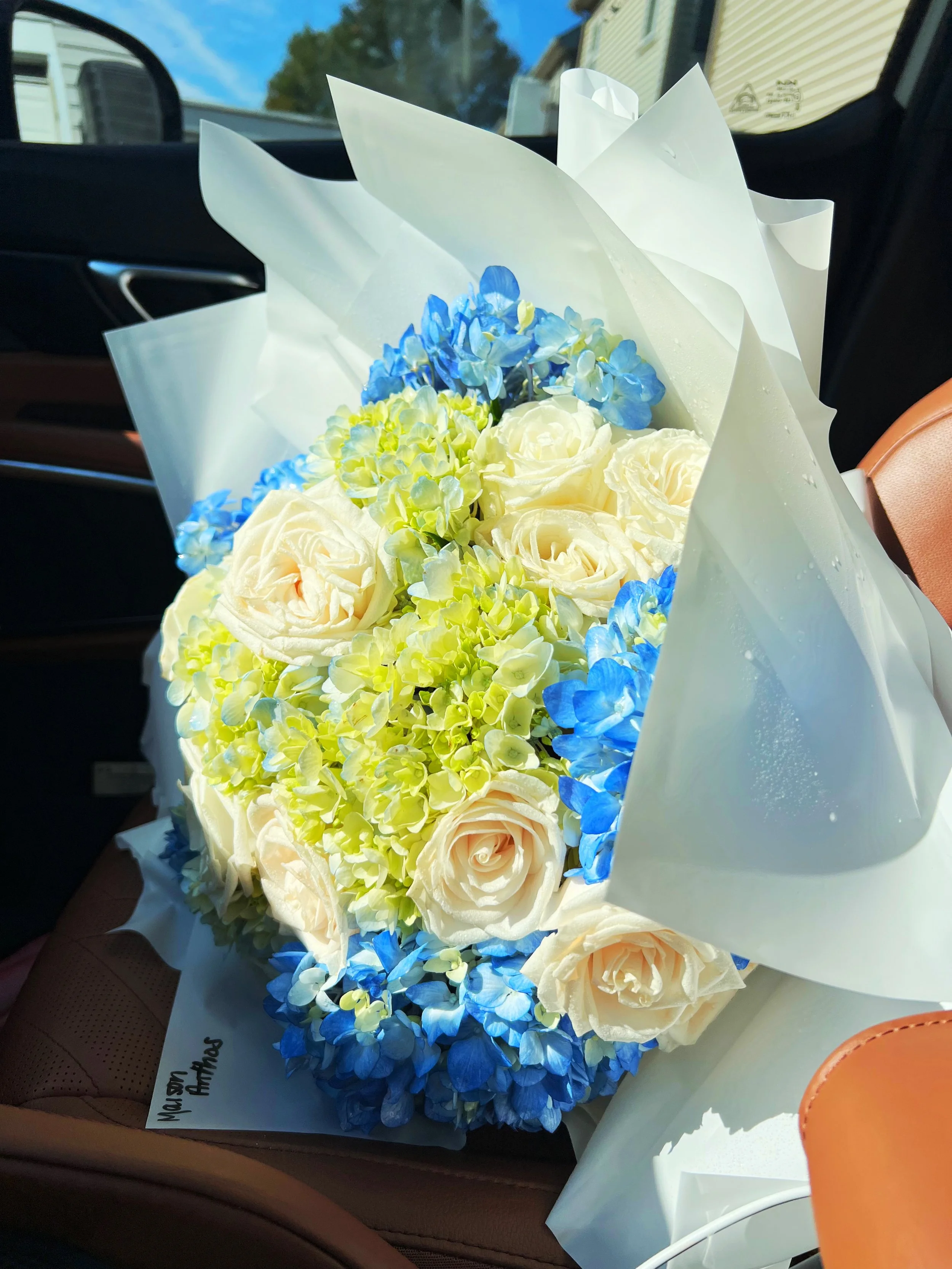A bouquet of white roses, blue and green hydrangeas wrapped in white paper, placed on the seat of a car.