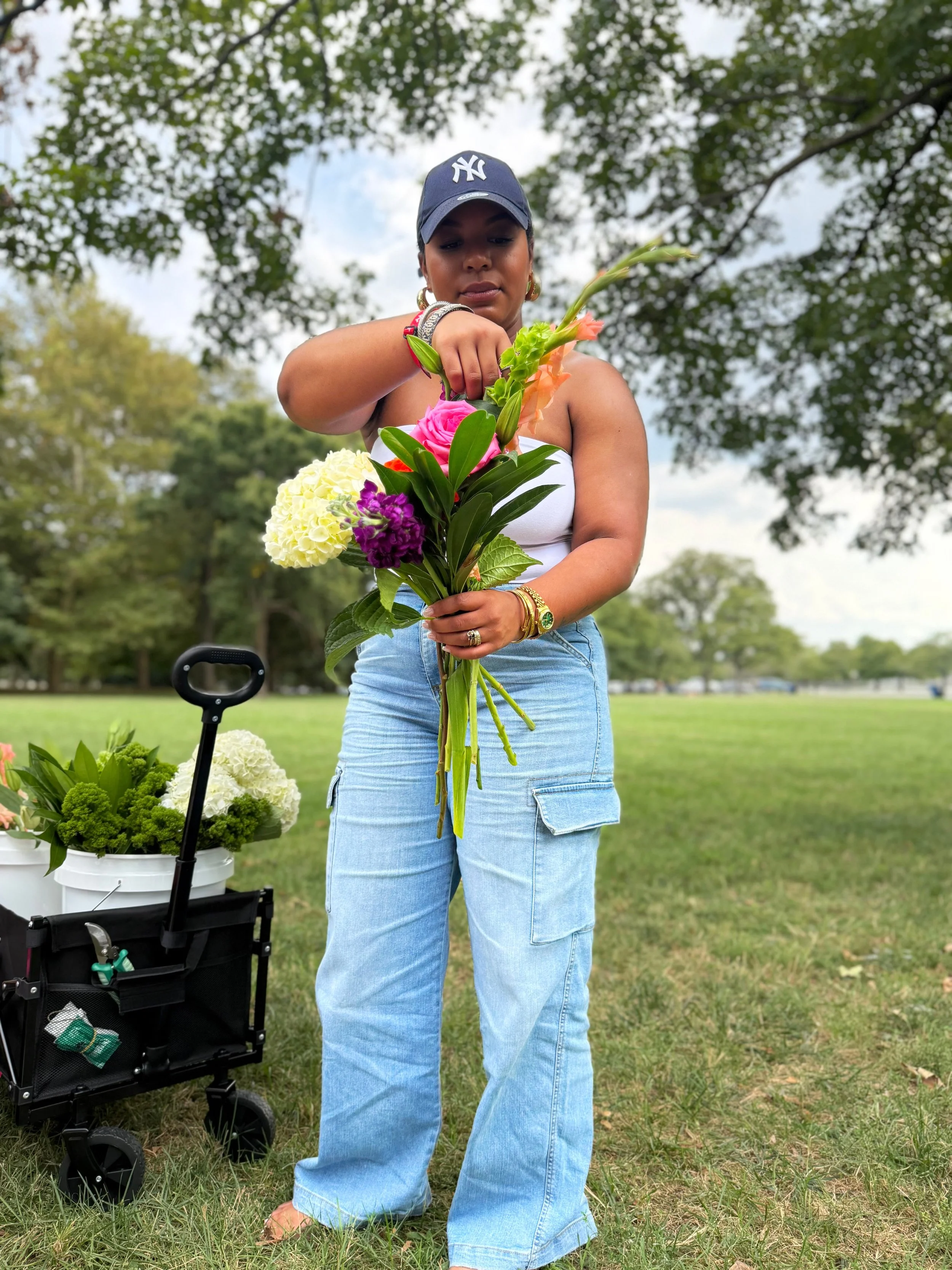 A woman wearing a navy baseball cap, white tank top, and light blue jeans arranges a bouquet of colorful flowers outdoors with trees and a grassy field in the background.