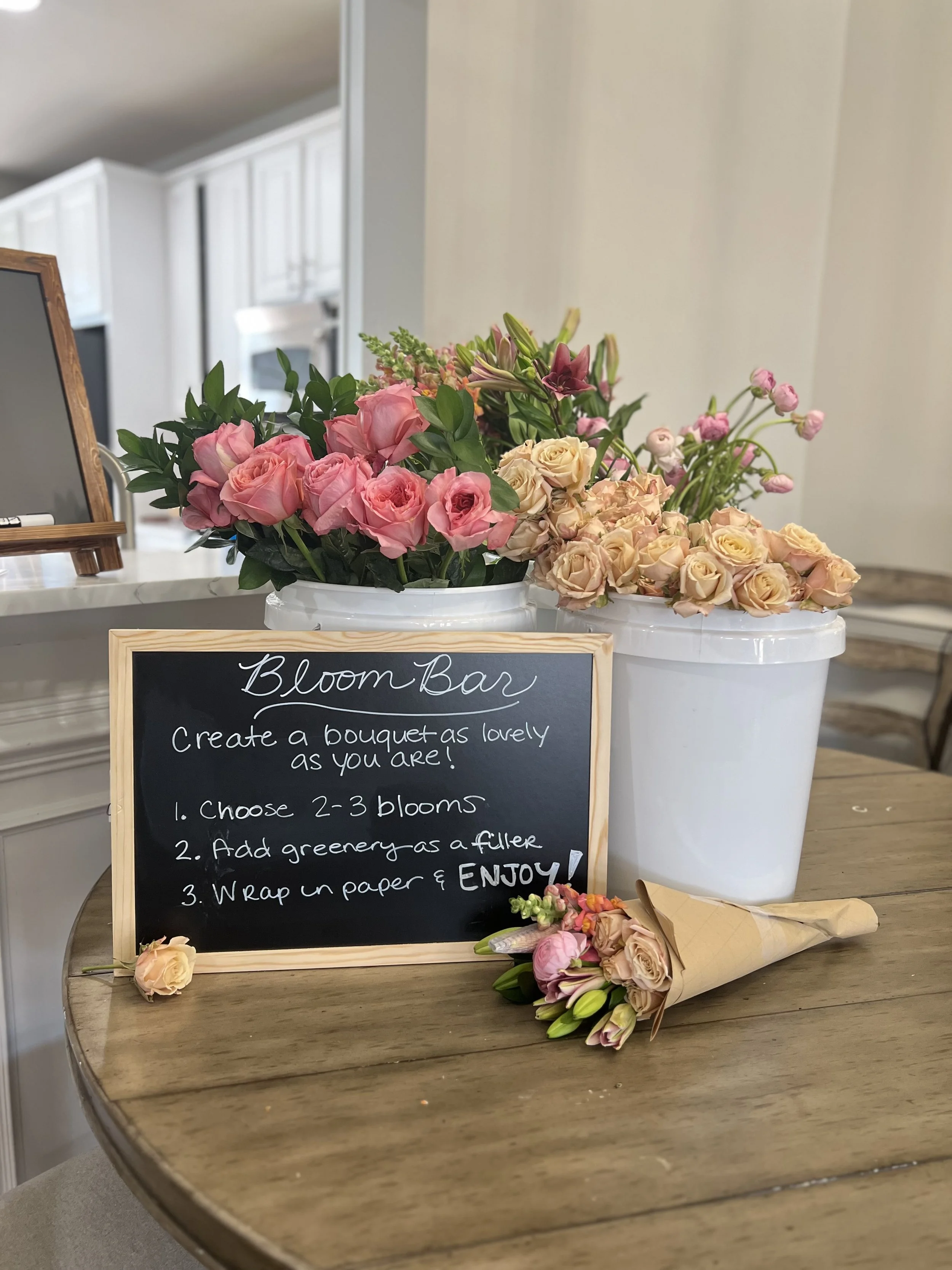 A floral arrangement on a wooden table with a chalkboard sign instructing how to create a flower bouquet, two large white pots with pink and peach roses, and a small bouquet of flowers wrapped in brown paper.