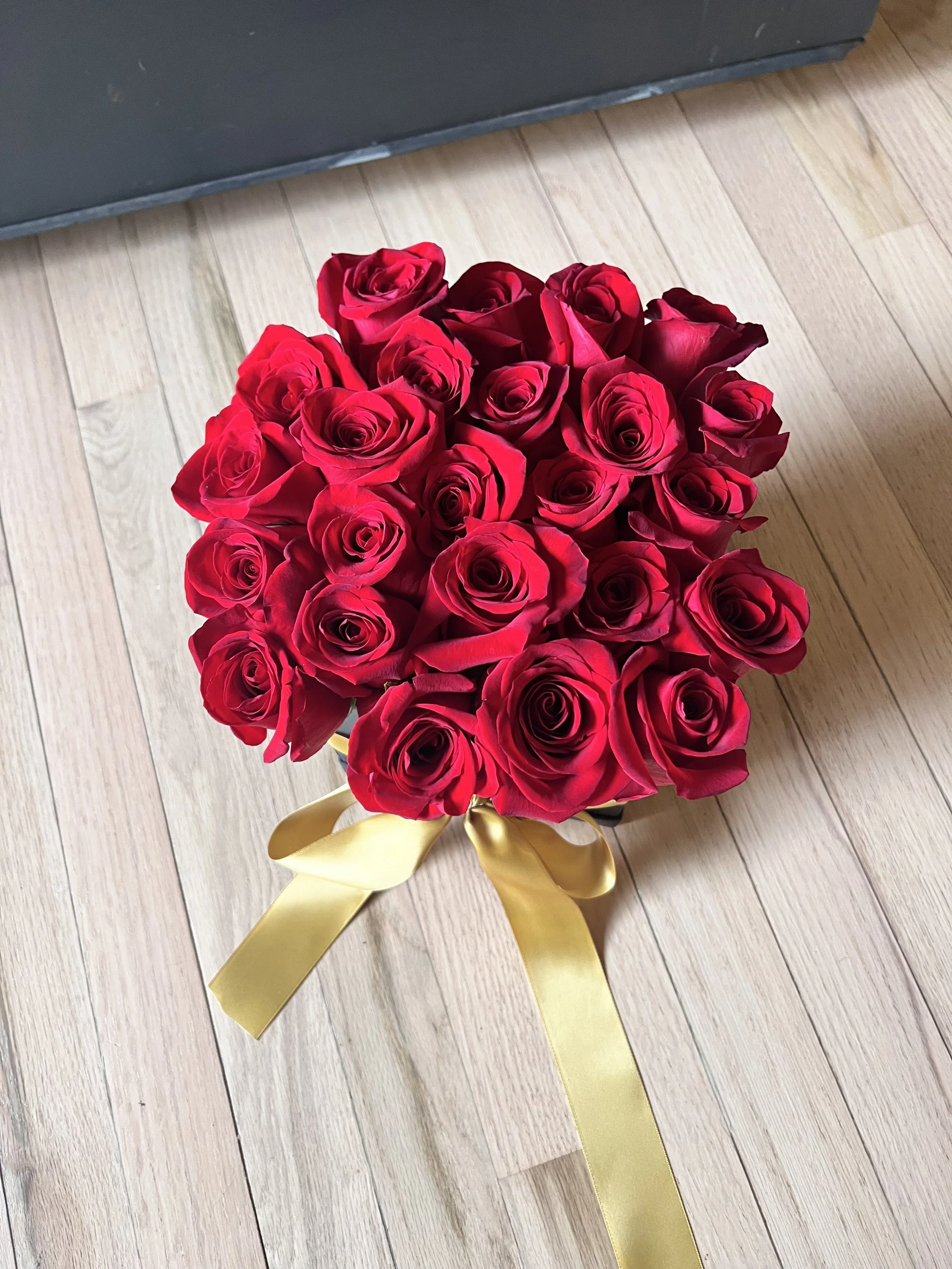 A bouquet of red roses tied with a gold ribbon placed on a light wooden floor.