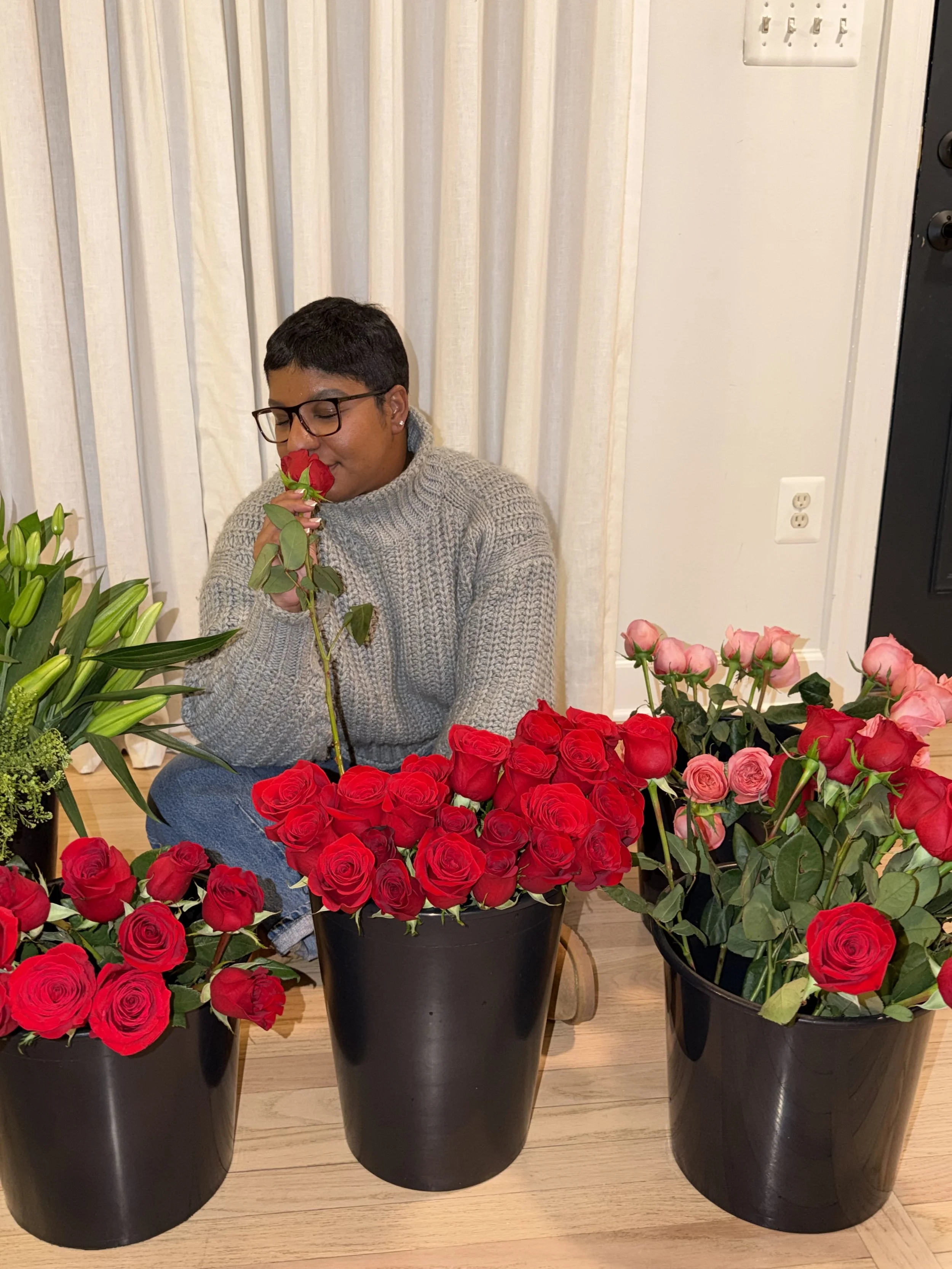 Person with short dark hair, glasses, and a gray sweater smelling a red rose, surrounded by three large black pots filled with red and pink roses, indoors with cream-colored curtains and a wooden floor.