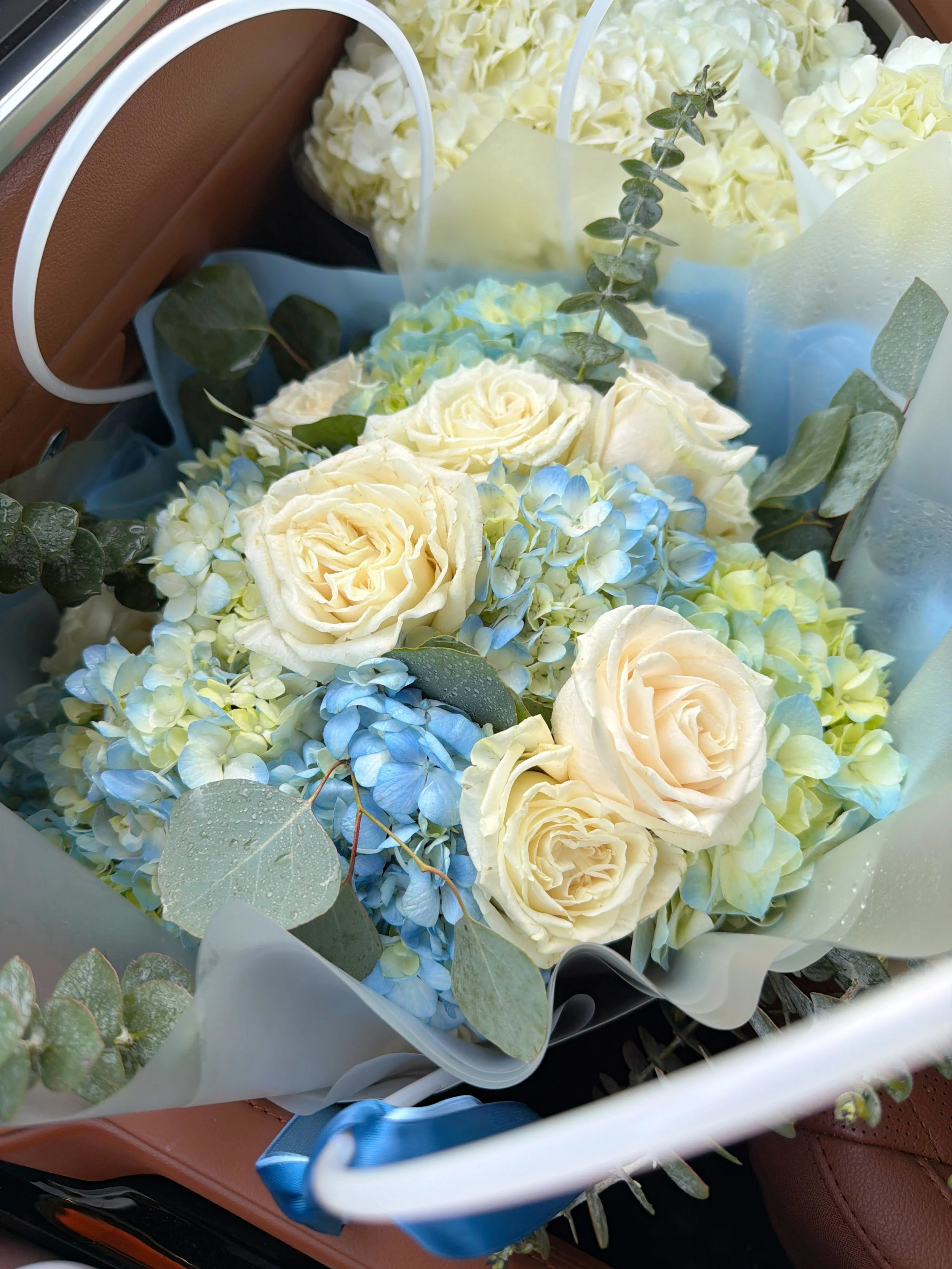 A bouquet of white roses, light blue hydrangeas, and greenery wrapped in light paper inside a white basket.