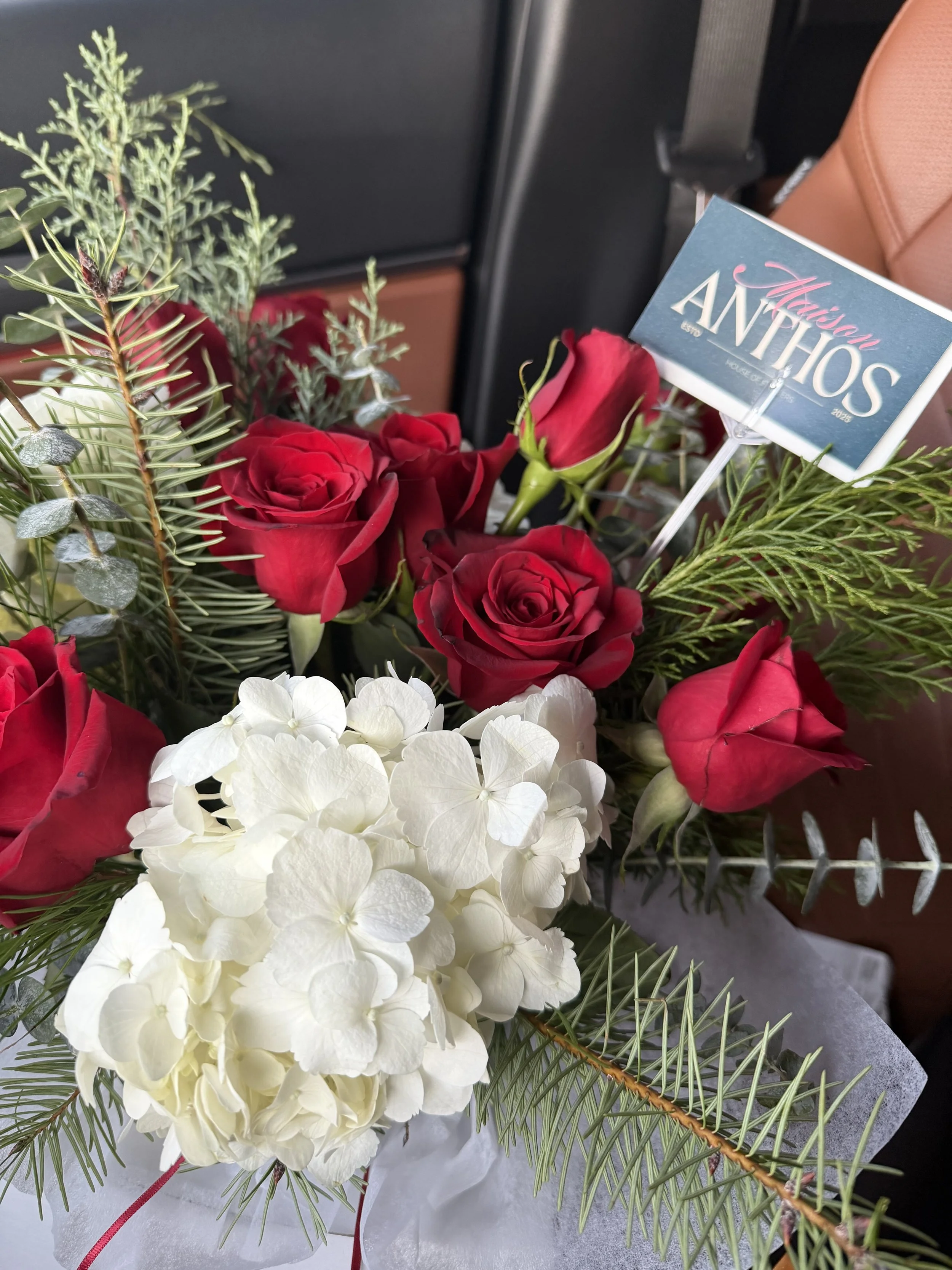 A floral arrangement with white hydrangeas and red roses surrounded by green fern leaves, placed on a surface in what appears to be a vehicle, with a card that reads "Missouri ANTHOS" inserted into the arrangement.