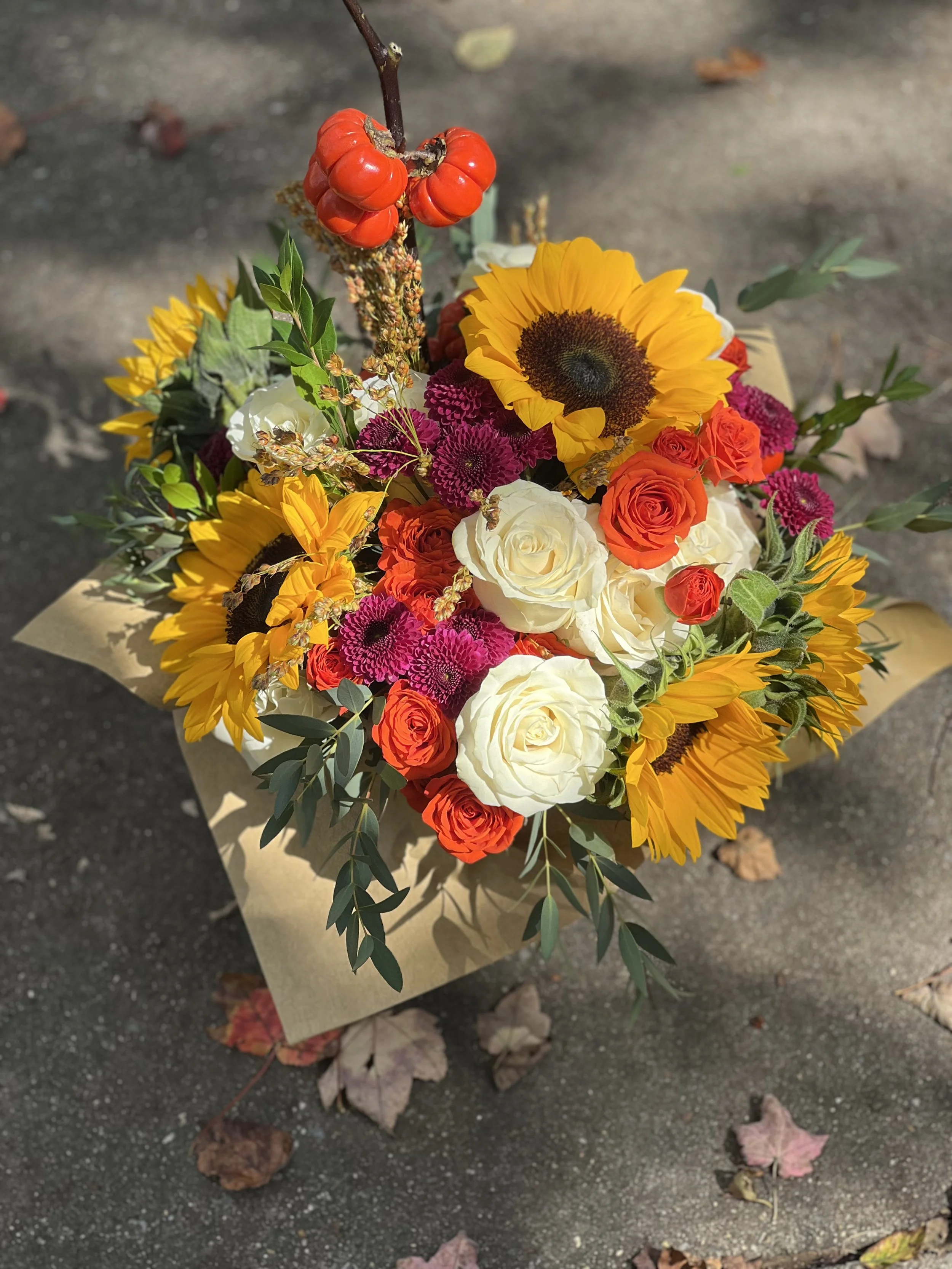 Colorful bouquet of sunflowers, white roses, orange roses, pink and purple flowers, with a red plant at the top, on brown wrapping paper on a pavement with fallen leaves.