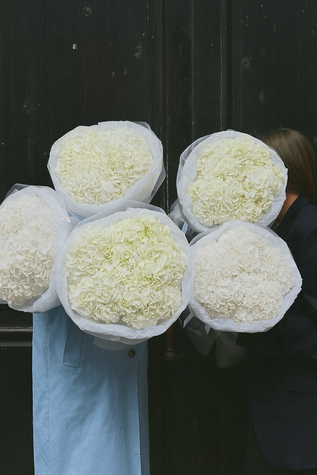 People holding large bouquets of white flowers, possibly roses or hydrangeas, wrapped in white paper, standing against a black wall.