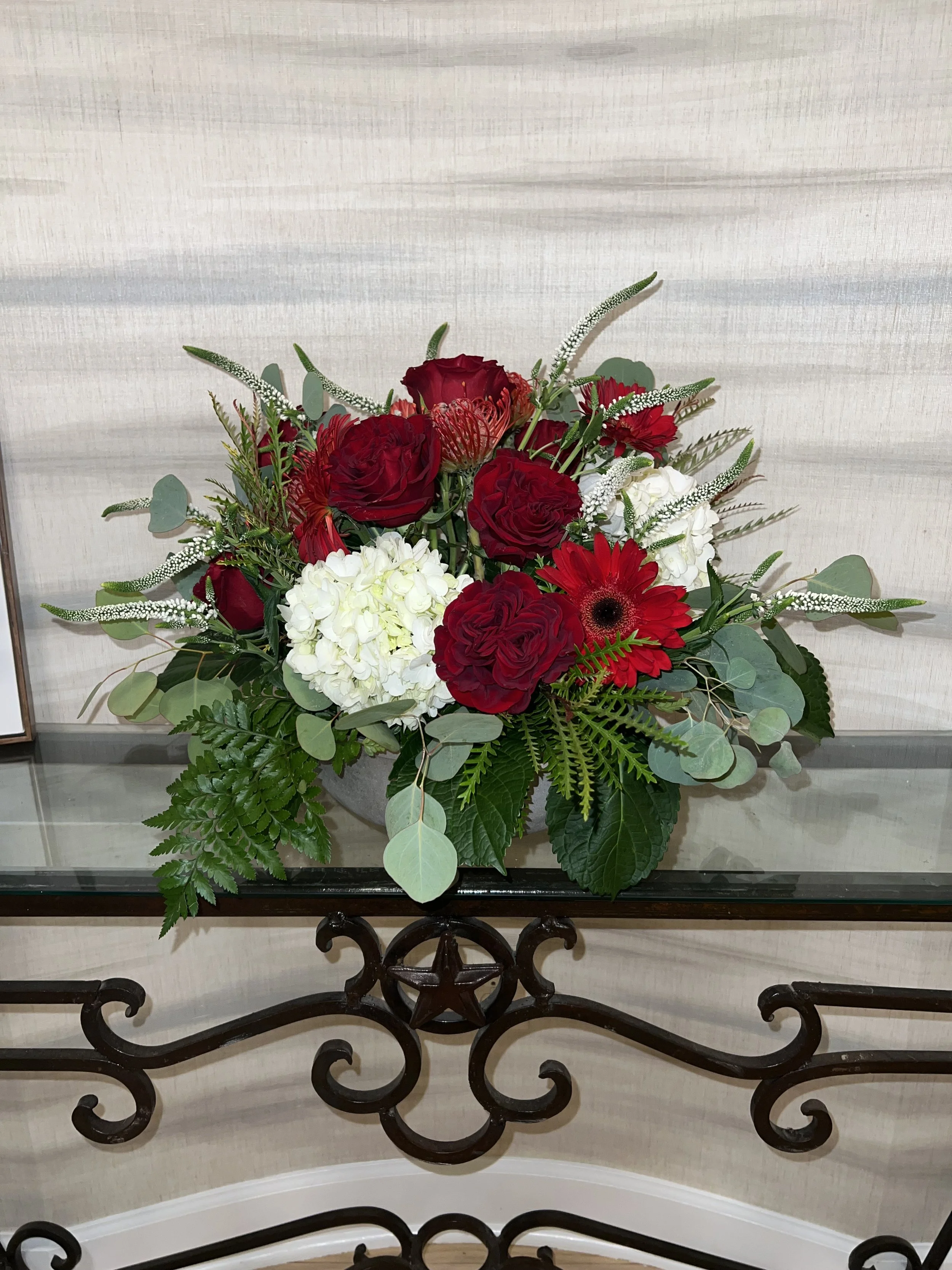 A floral arrangement with red roses, white hydrangeas, red gerbera daisies, red matthiolas, and greenery, displayed on a glass table with a decorative metal base.