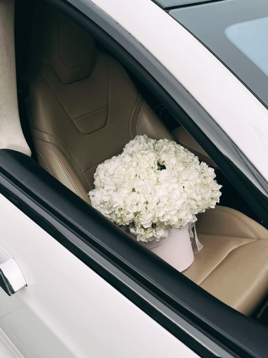 A white luxury car with a large bouquet of white hydrangea flowers on the beige leather seat visible through the open sunroof.
