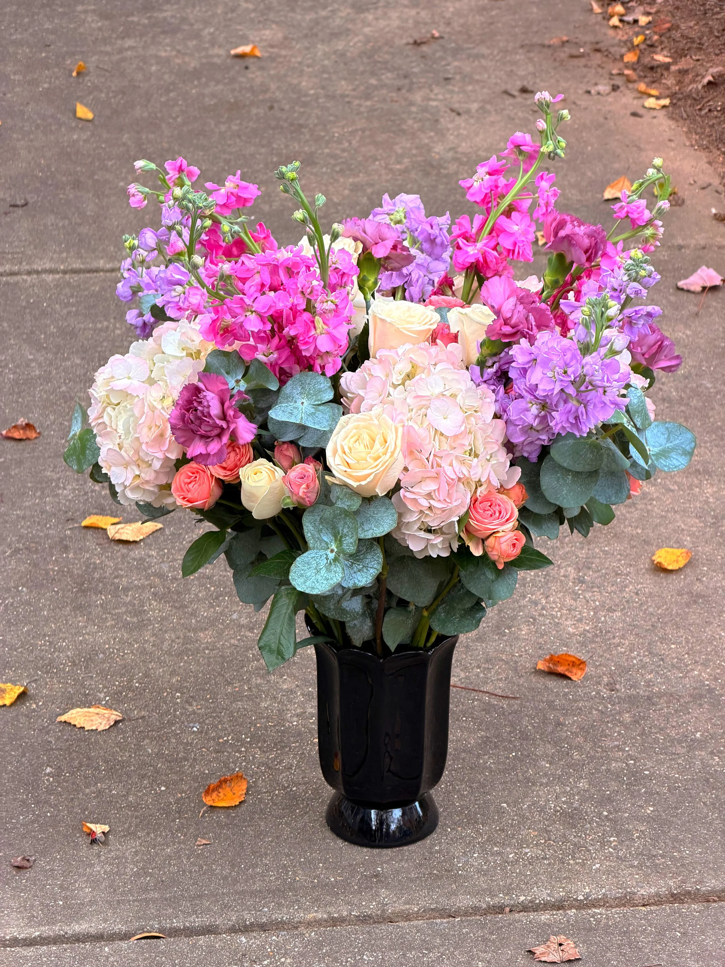 A bouquet of colorful flowers in a black vase on a concrete sidewalk with fallen leaves.