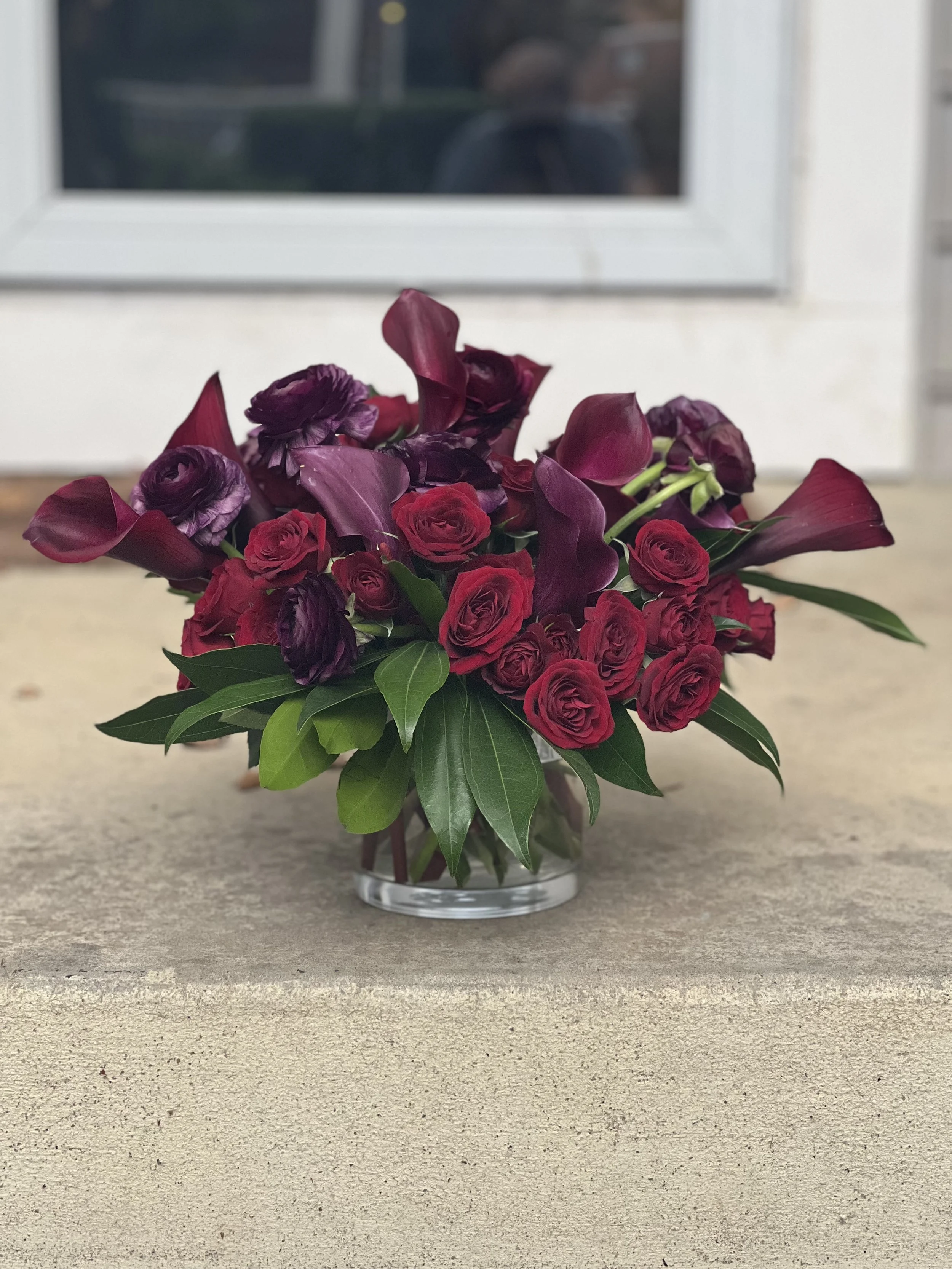 A floral arrangement with red roses, purple calla lilies, and green foliage in a clear glass vase on a concrete surface outside a house with a window in the background.