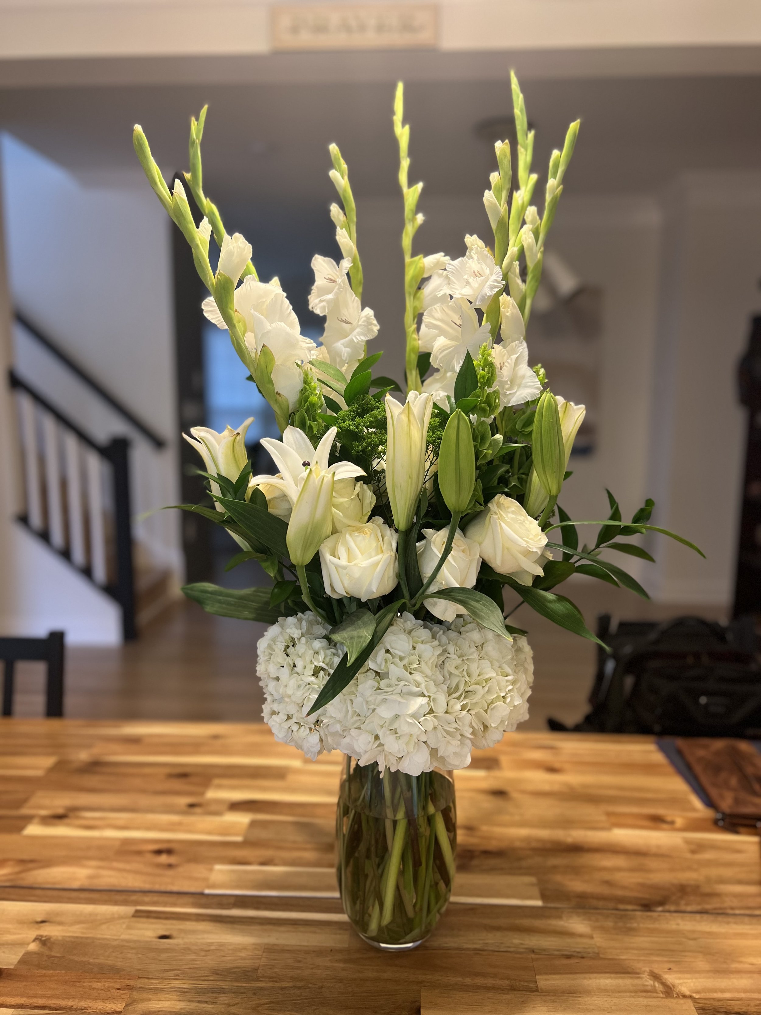 A tall glass vase filled with white flowers including gladiolus, roses, lilies, and hydrangeas on a wooden table in a cozy indoor setting.