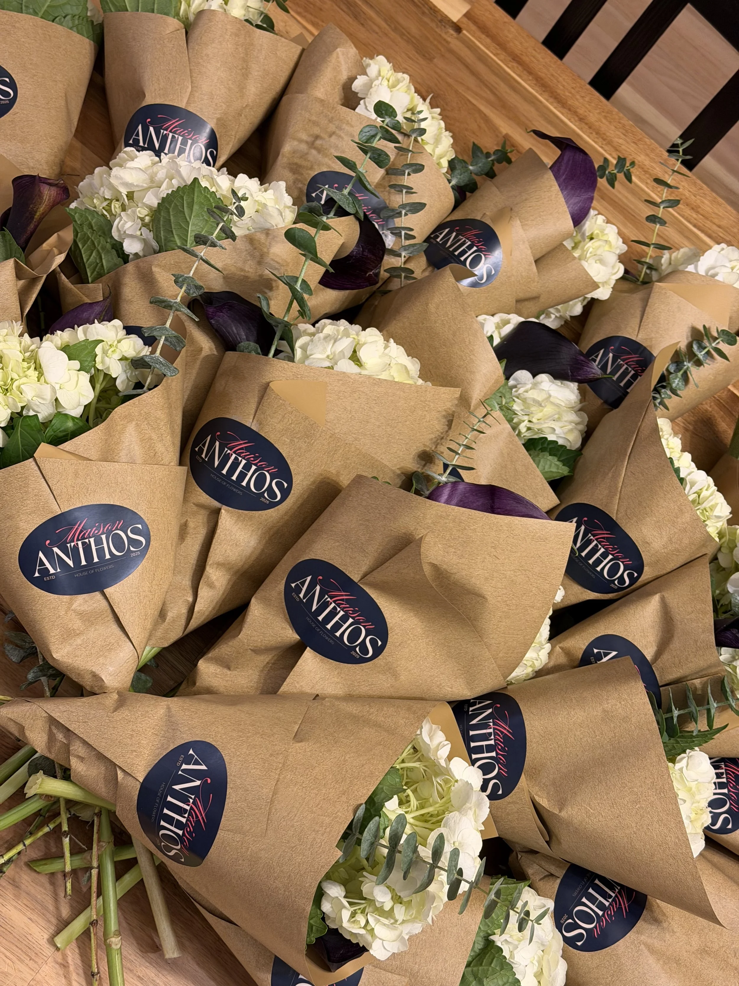 Multiple bouquets of white hydrangeas and dark purple calla lilies wrapped in brown paper with 'Maison Anthos' stickers, arranged on a wooden table.