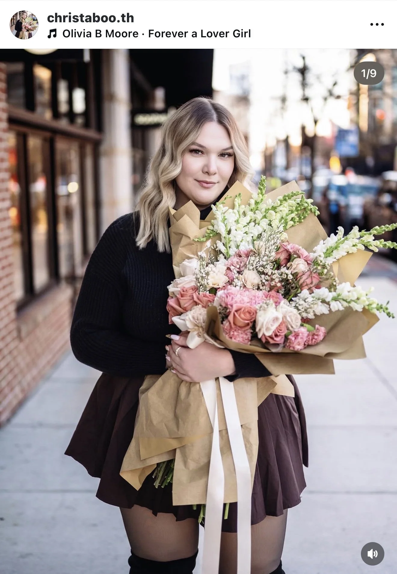 A young woman with shoulder-length blonde hair wearing a black sweater and a dark skirt, standing on a city sidewalk while holding a large bouquet of pink, white, and peach flowers wrapped in brown paper.