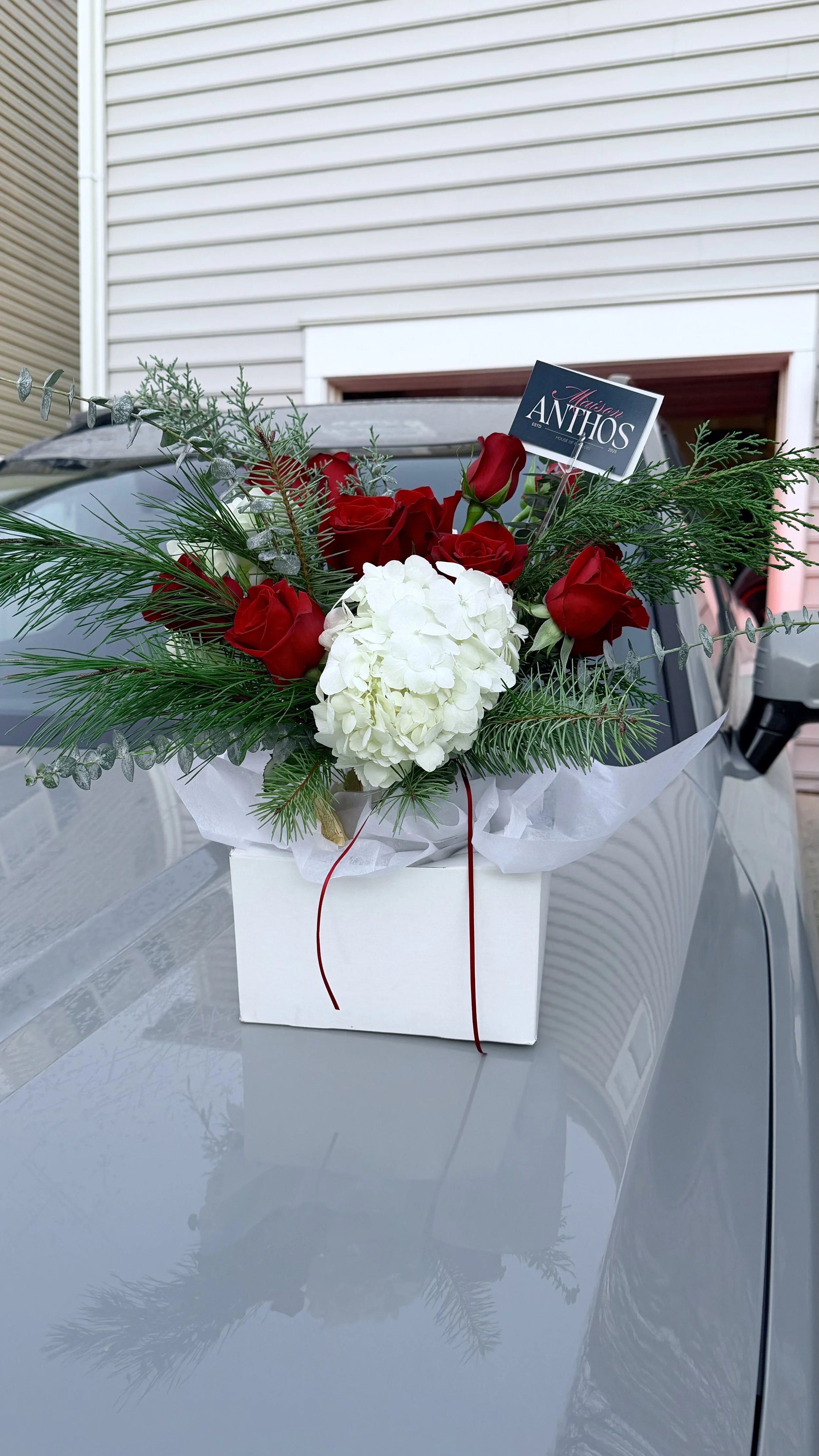A floral arrangement with red roses, white hydrangeas, and greenery in a white box, placed on a silver car hood, with a house and garage in the background.